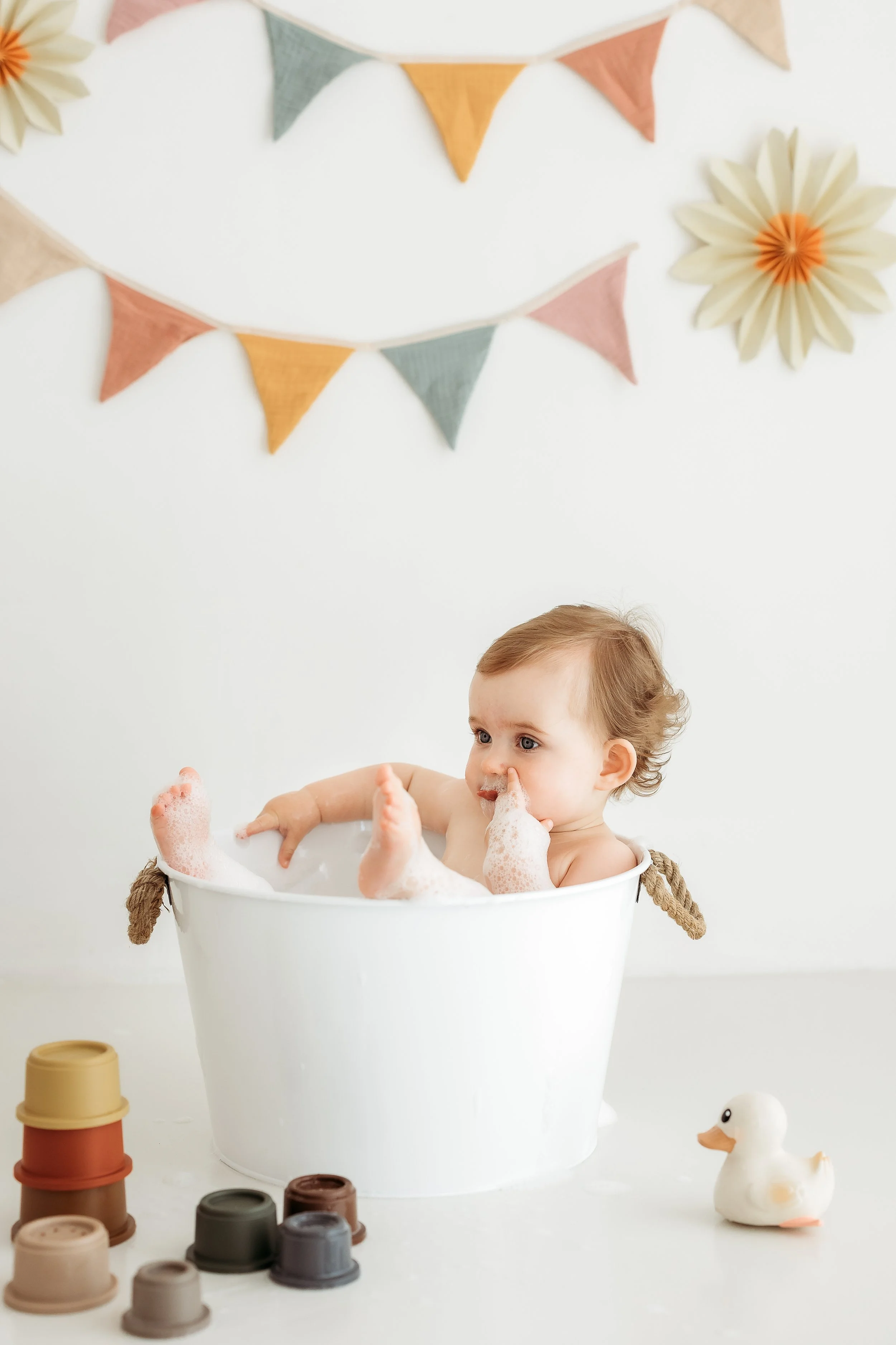 One year old child enjoying a bubble bath in a white tub during a playful first birthday photography session