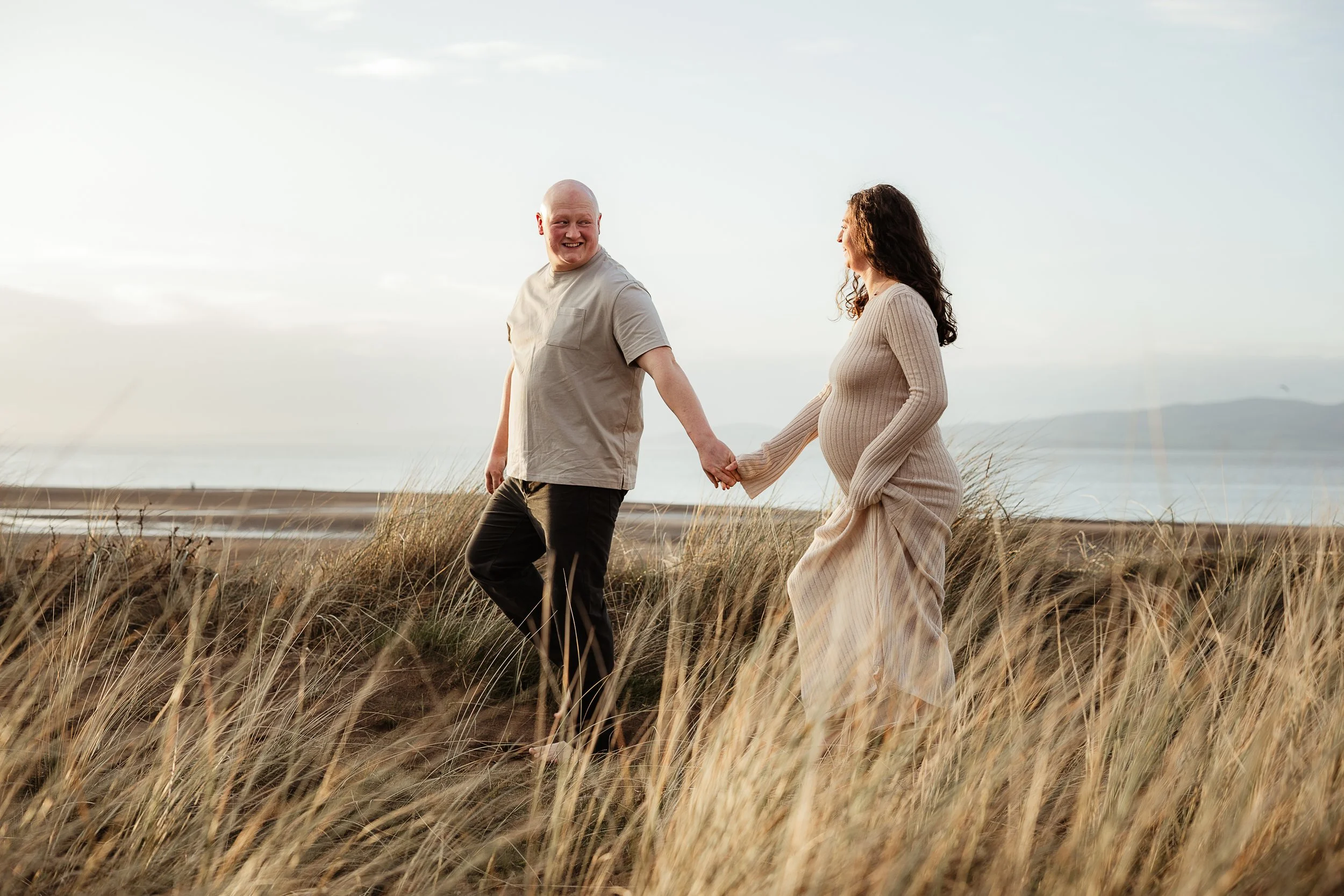 Expectant couple walking hand in hand through long grass during an outdoor maternity session.