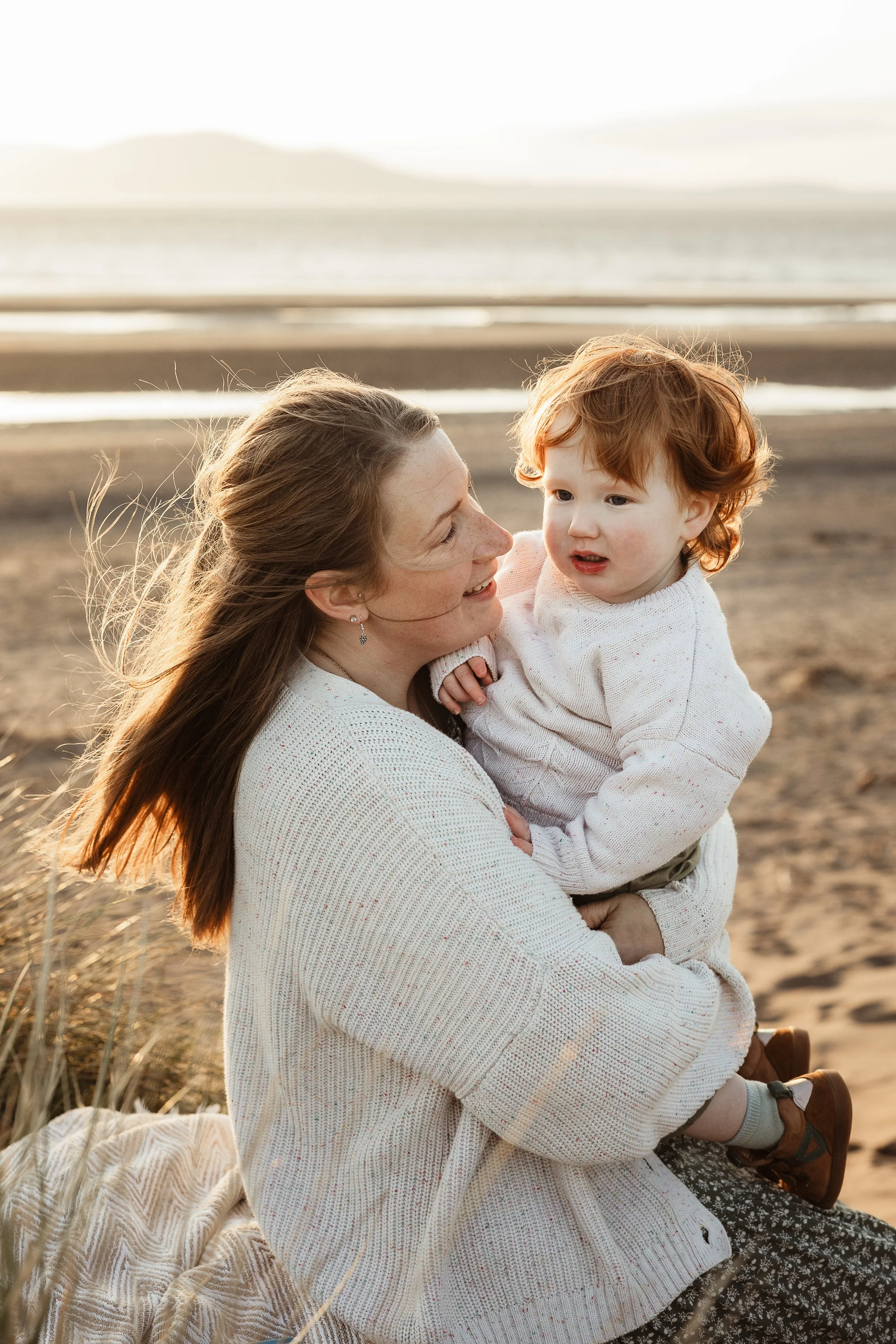 Mother and young child walking together along the beach at sunset, with the ocean and mountains in the background.