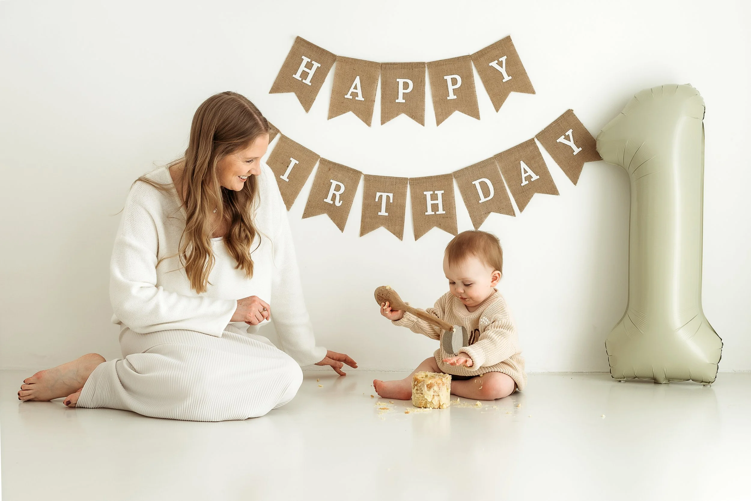 Mother and child celebrating a first birthday together with cake, balloons and birthday decorations in a natural light studio