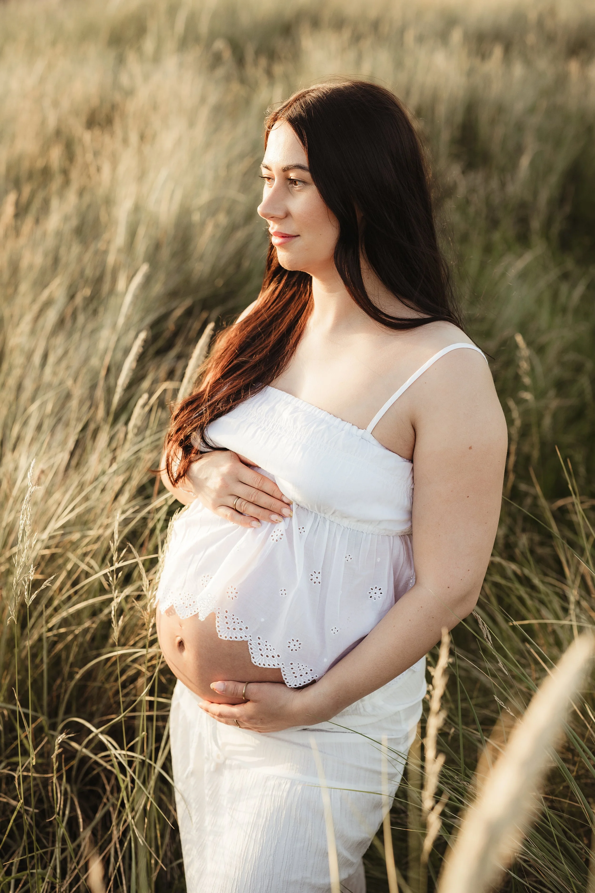 A mother-to-be surrounded by natural light and tall grasses during a relaxed outdoor maternity photography session.