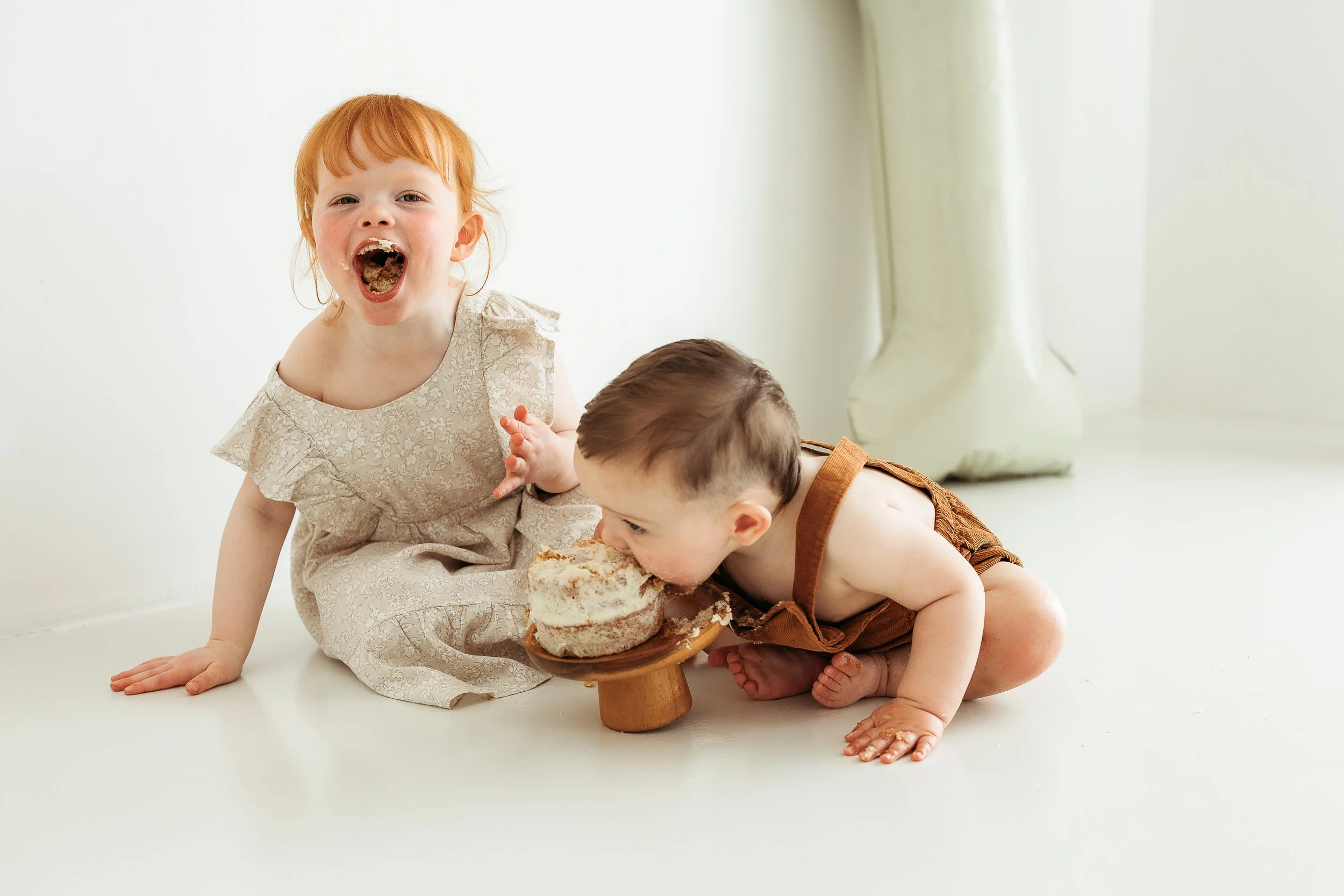 Two young children sitting together enjoying cake during a fun and messy first birthday photography session.