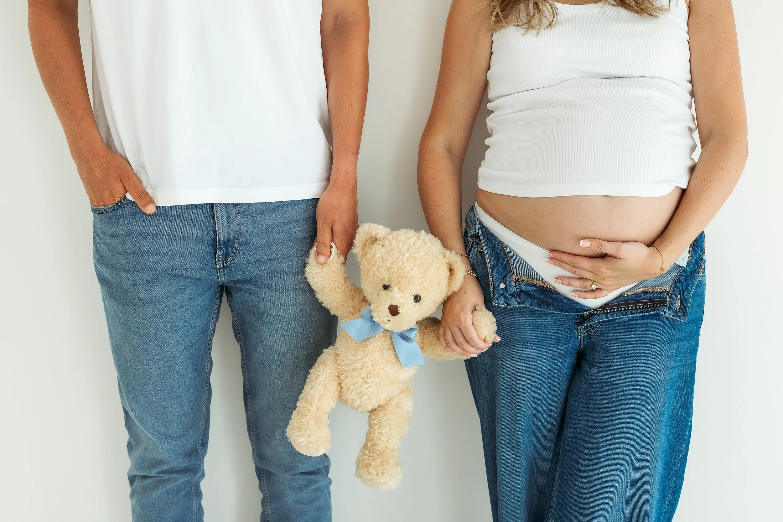 Expectant parents holding a gender reveal teddy bear with a blue ribbon and cradling the baby bump during a studio maternity session.