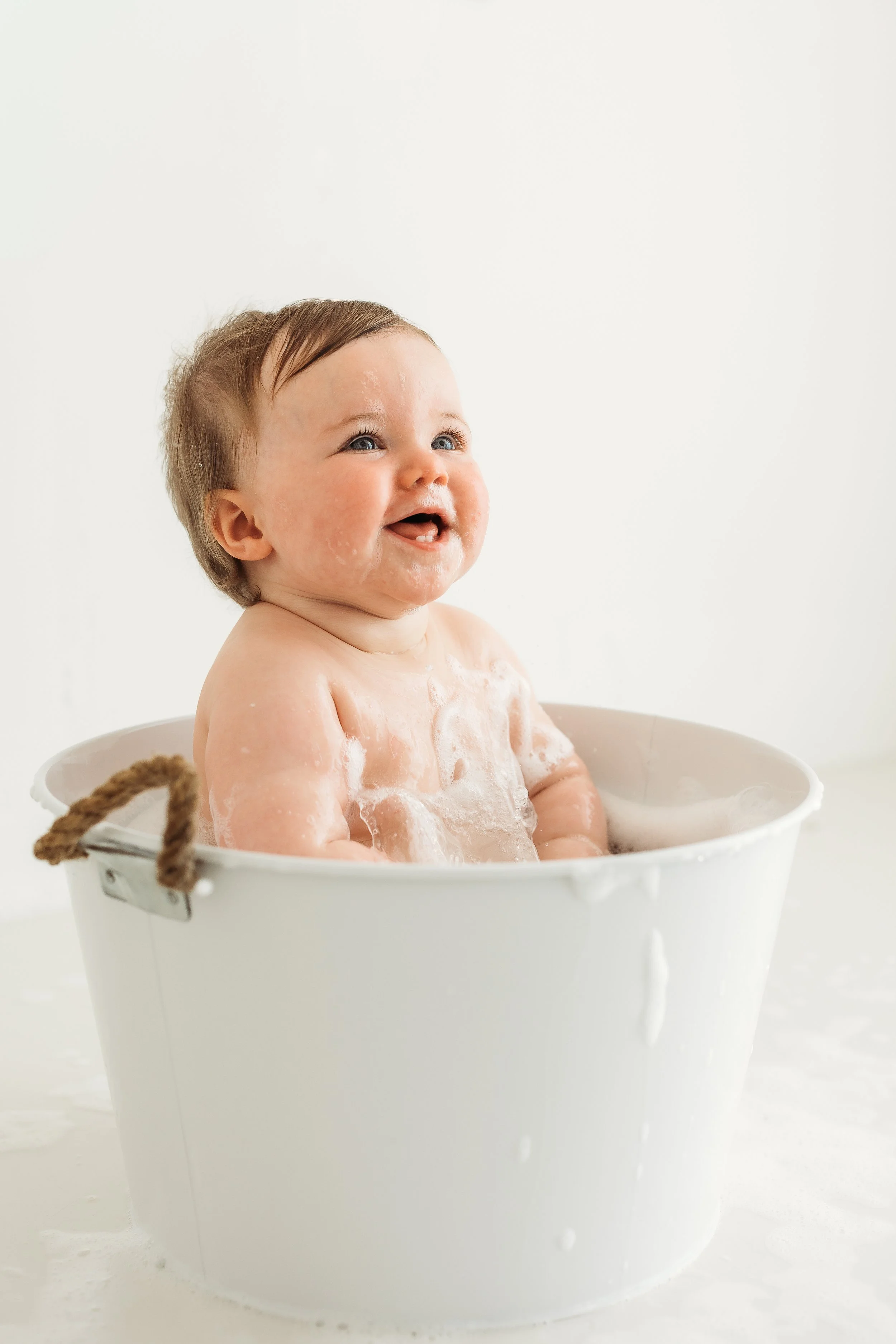 Smiling baby sitting in a white tub surrounded by bubbles during a relaxed first birthday photography session