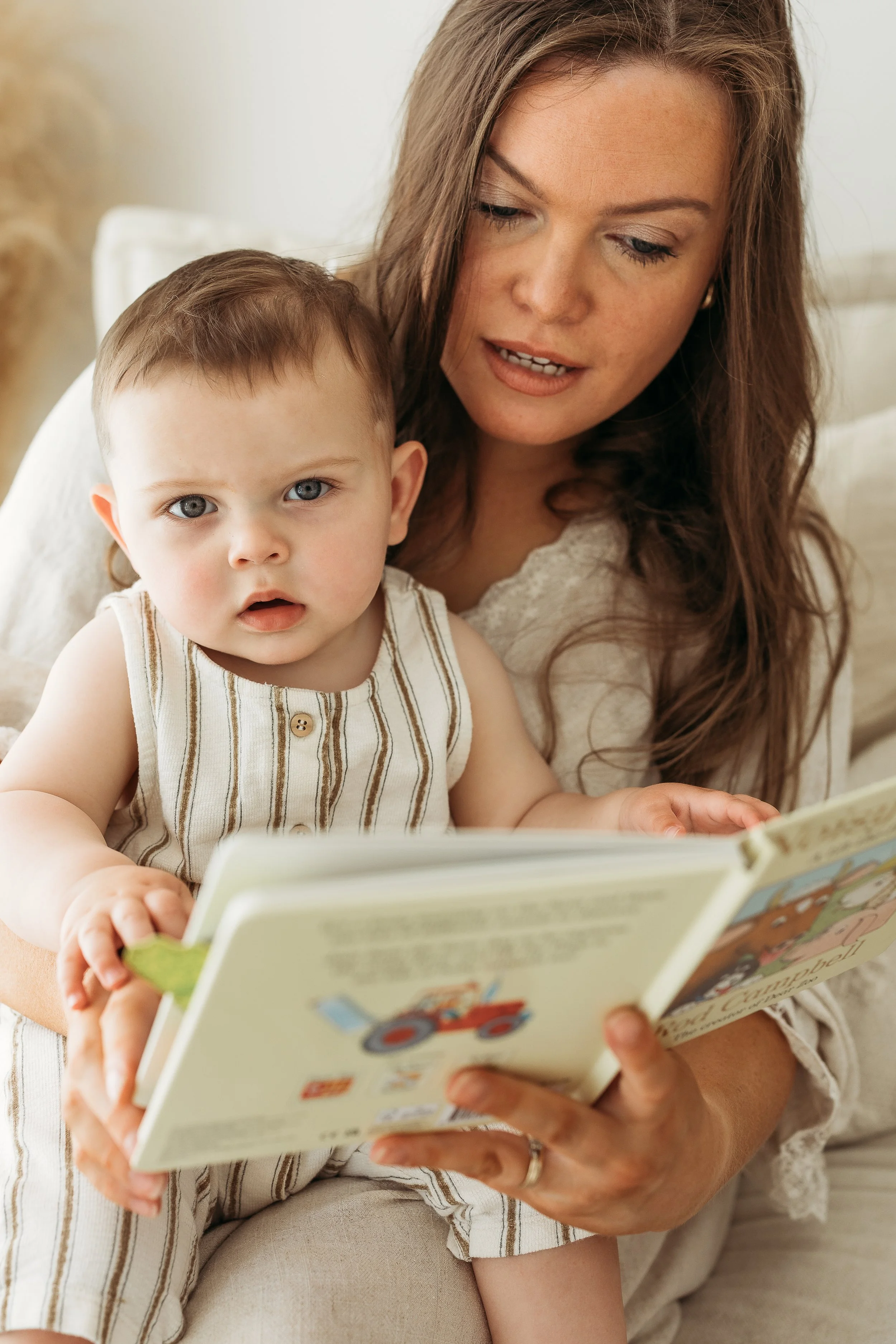 Mother reading a story book with her young son on a beige sofa during a relaxed first birthday photography session.