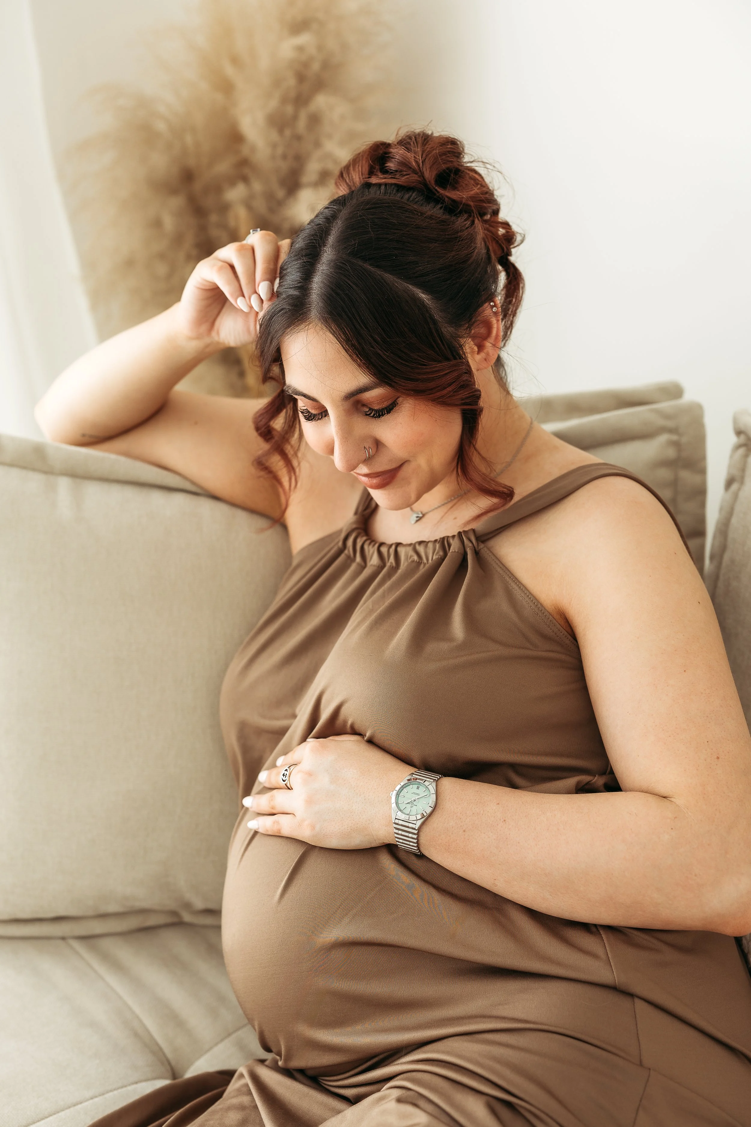 Pregnant woman sitting on a sofa with her hands resting on her bump during a relaxed maternity session.