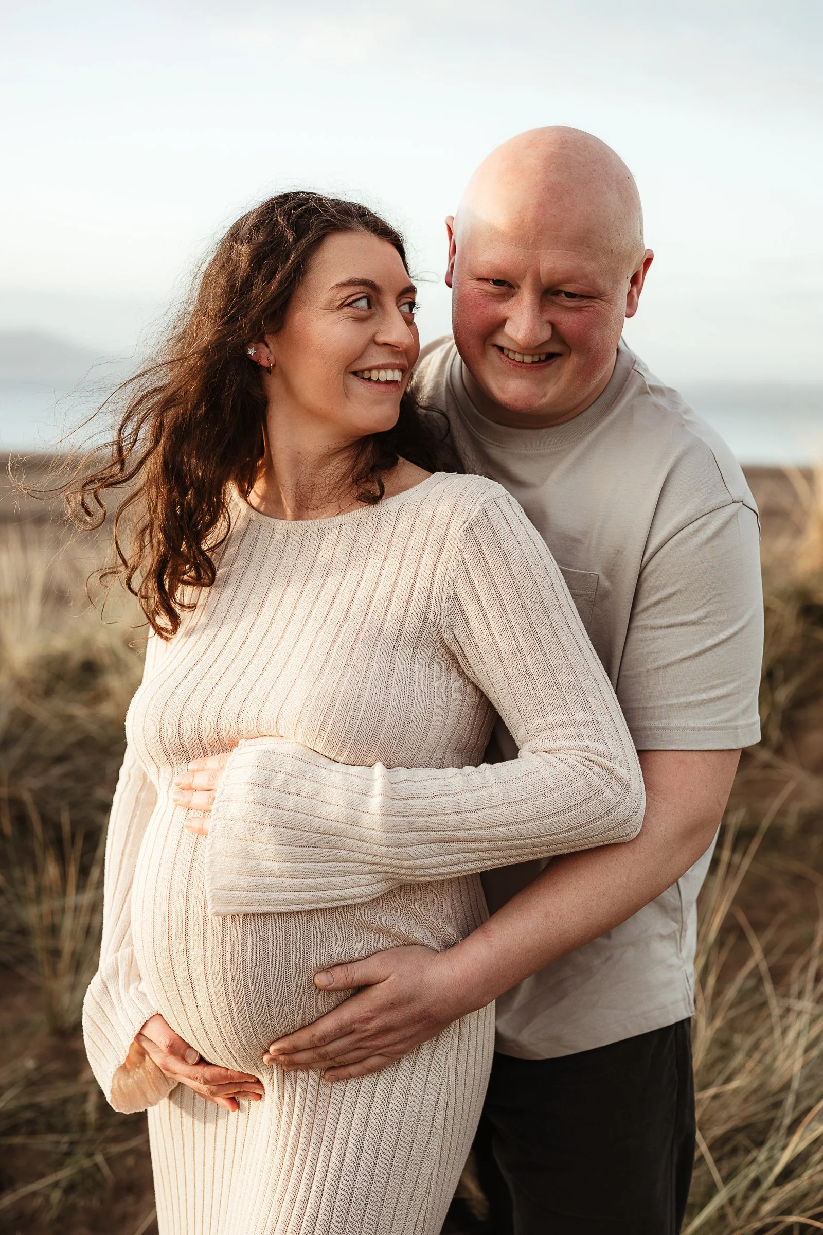 Expecting parents embracing during an outdoor maternity photography session by the coast.