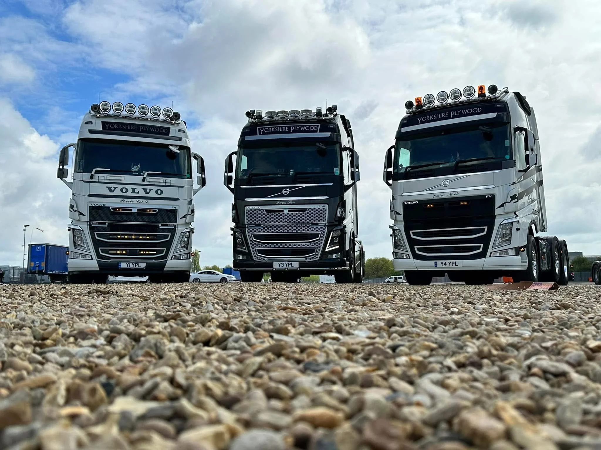 Three large trucks parked on gravel under a cloudy sky, with trees and a building in the background.