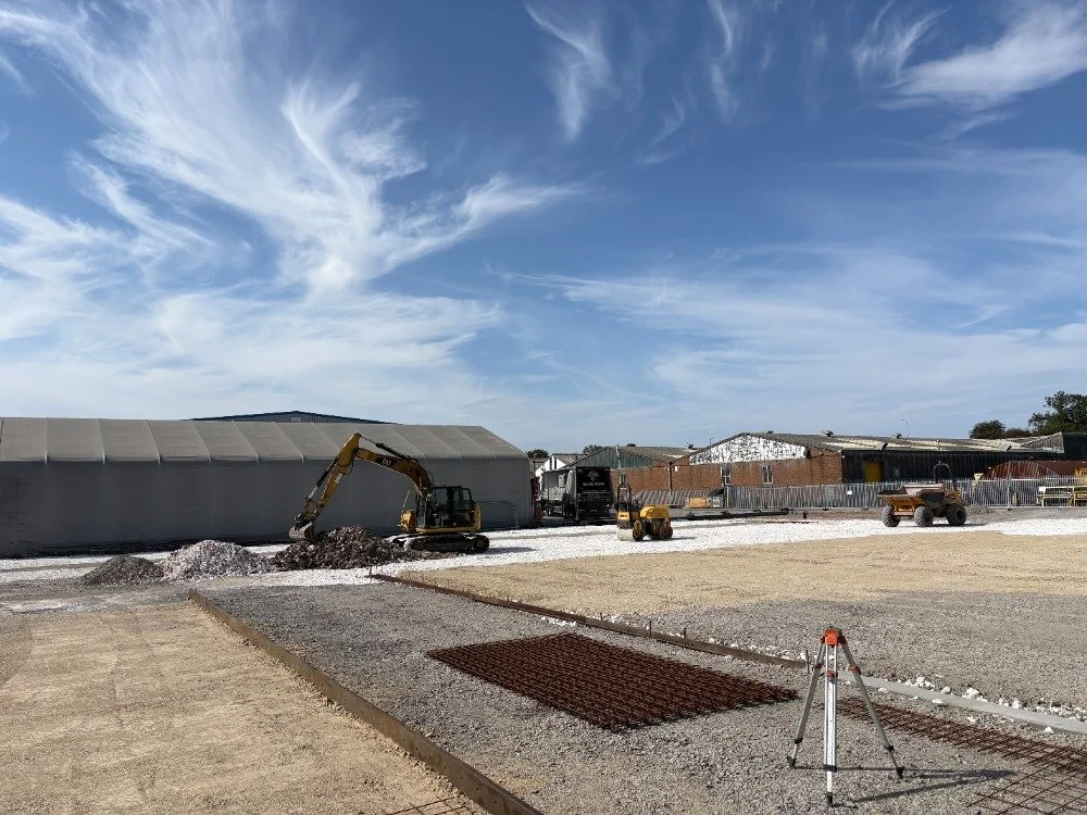 Construction site with small excavator digging gravel, barriers, construction equipment, and a large building structure in the background under a partly cloudy sky.