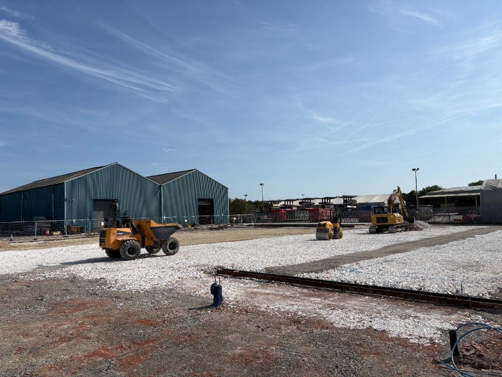 Construction site with heavy machinery, two green industrial buildings, and a gravel-covered area under a blue sky.