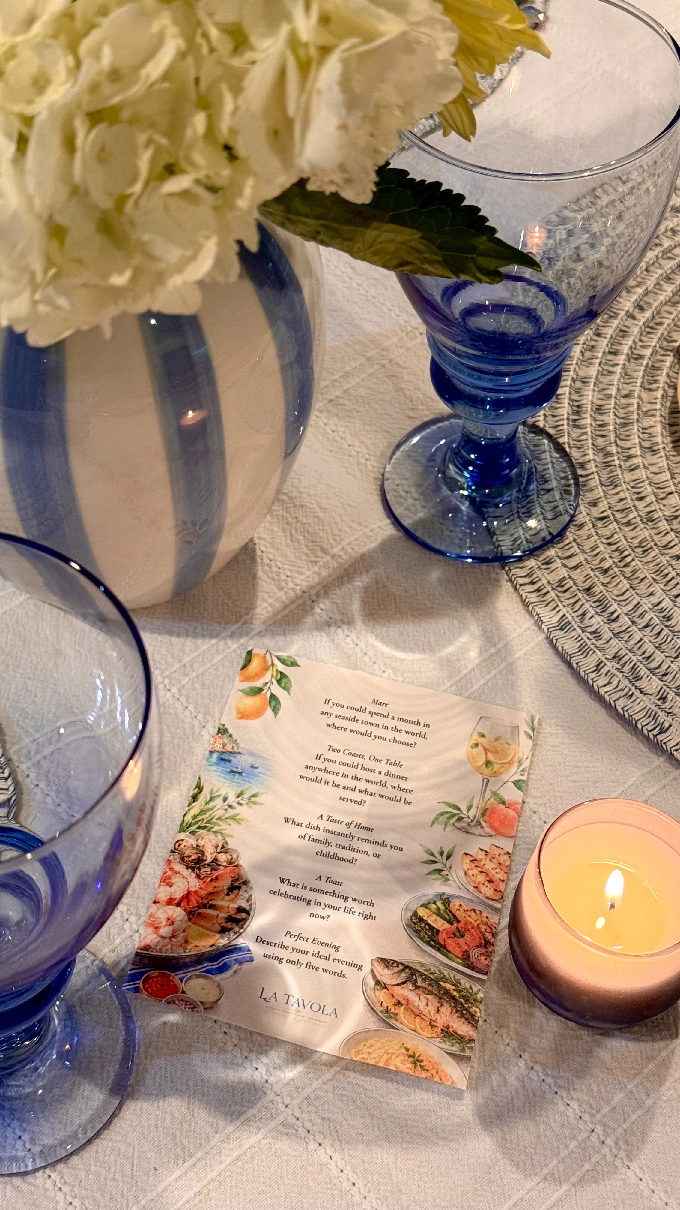 A table setting with a white tablecloth, a blue and white striped vase with white flowers, a blue glass stemmed glass, lit candle in a pink holder, and a menu with food illustrations.