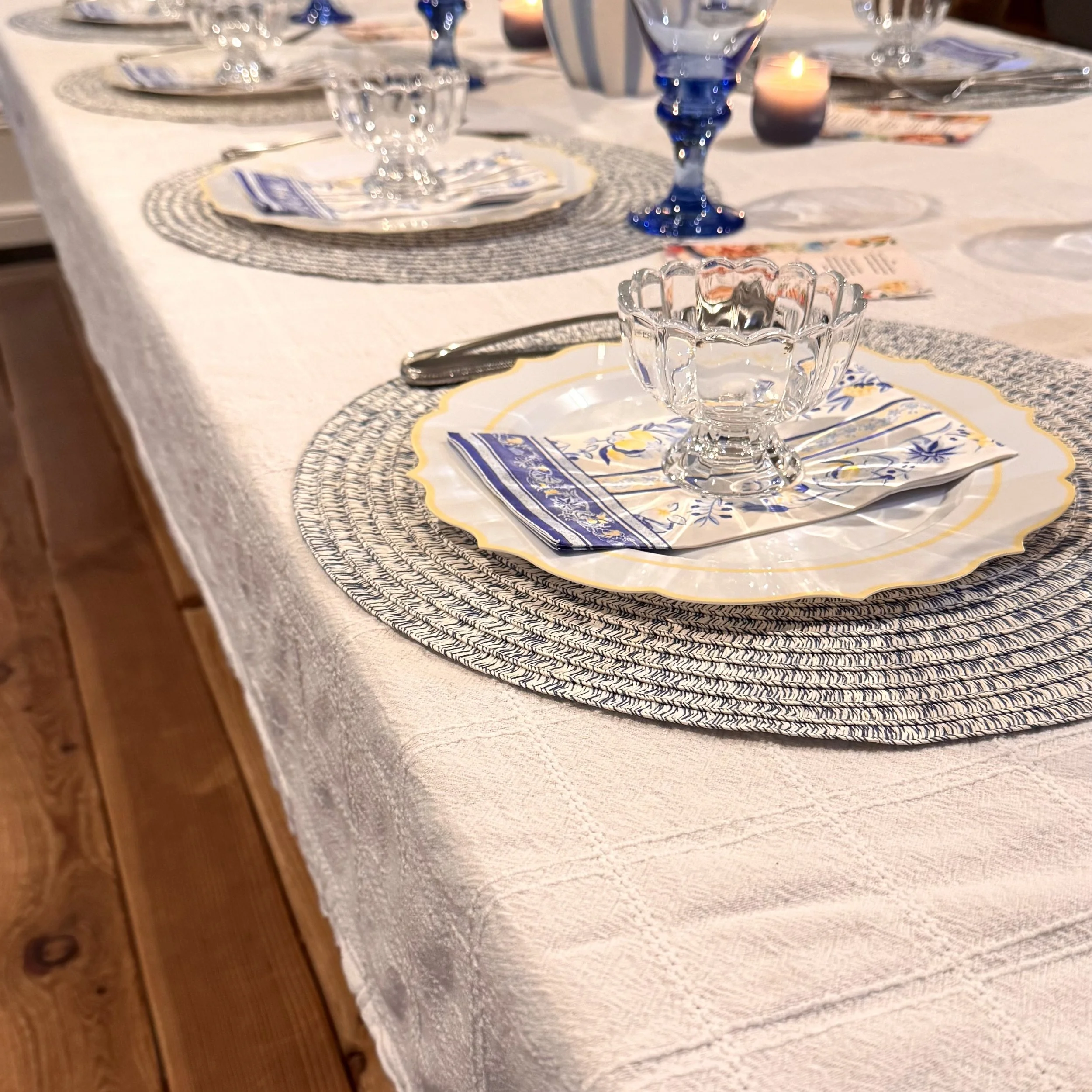 A dinner table setting with white tablecloth, decorative placemats, blue Goblets, an empty glass bowl, and a candle in the background.