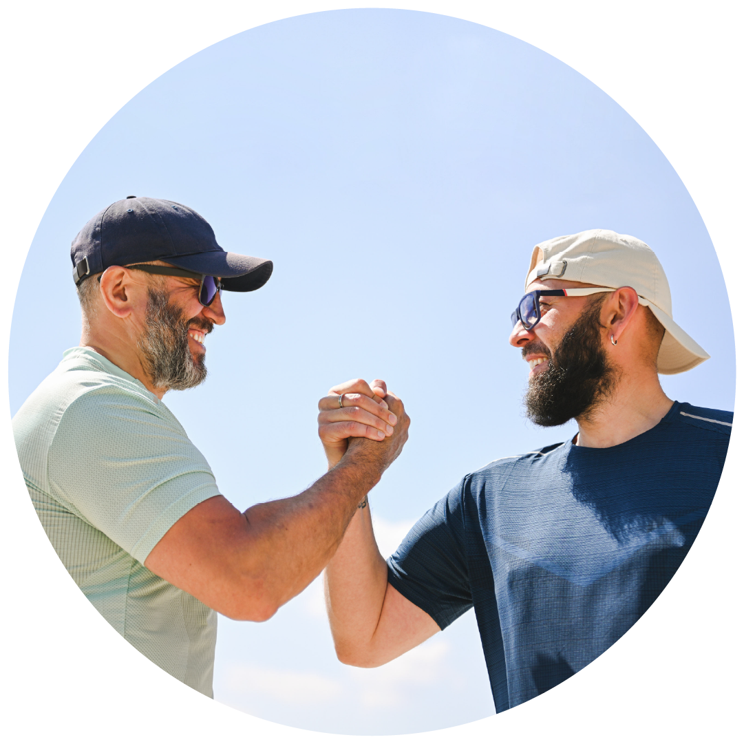 Two men with beards and sunglasses, wearing baseball caps, engaged in a friendly arm wrestling match outdoors against a clear blue sky.