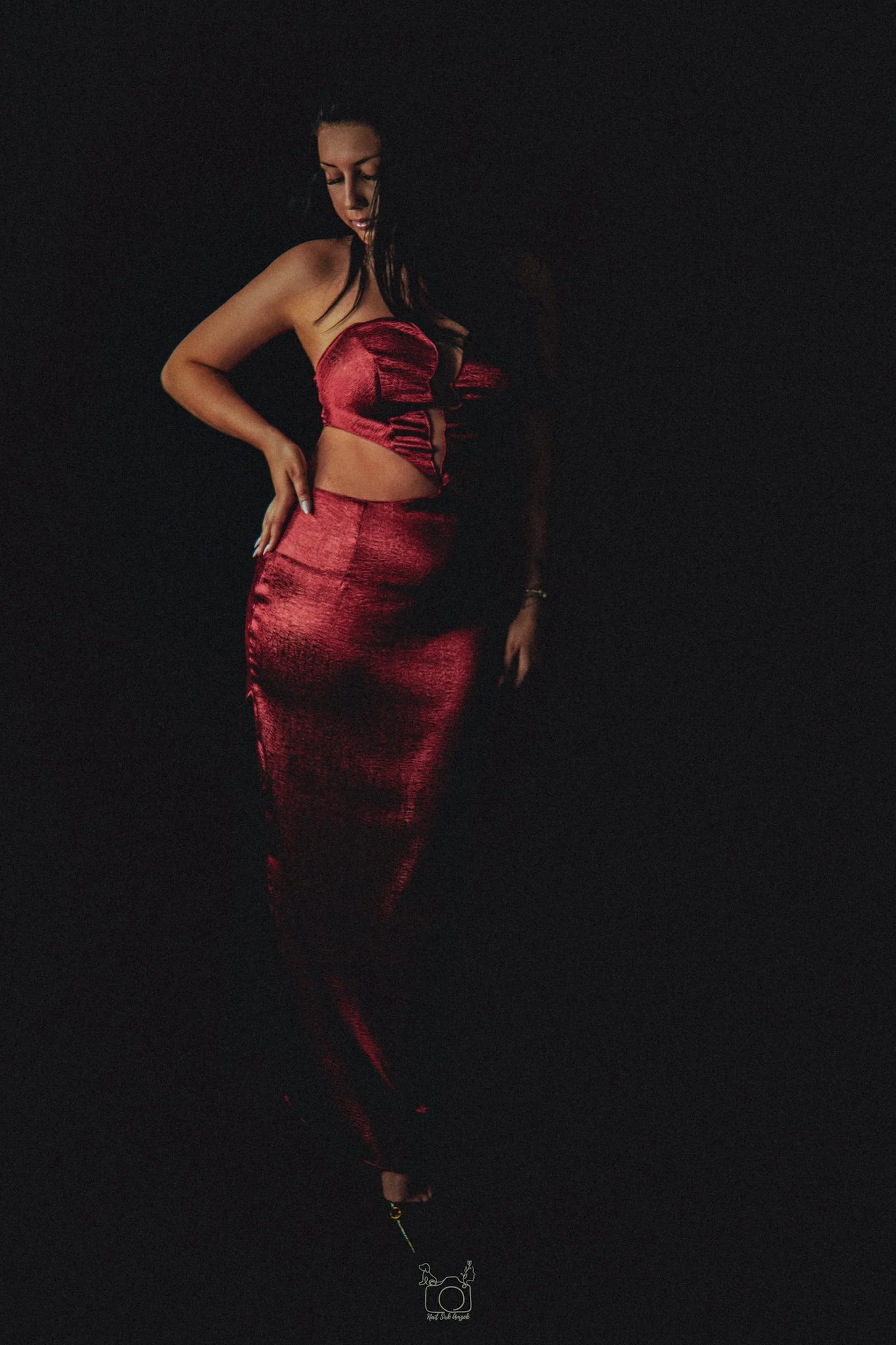 A woman in a strapless red satin dress standing in a dark background. She has long dark hair and appears to be looking down with her hand on her hip.