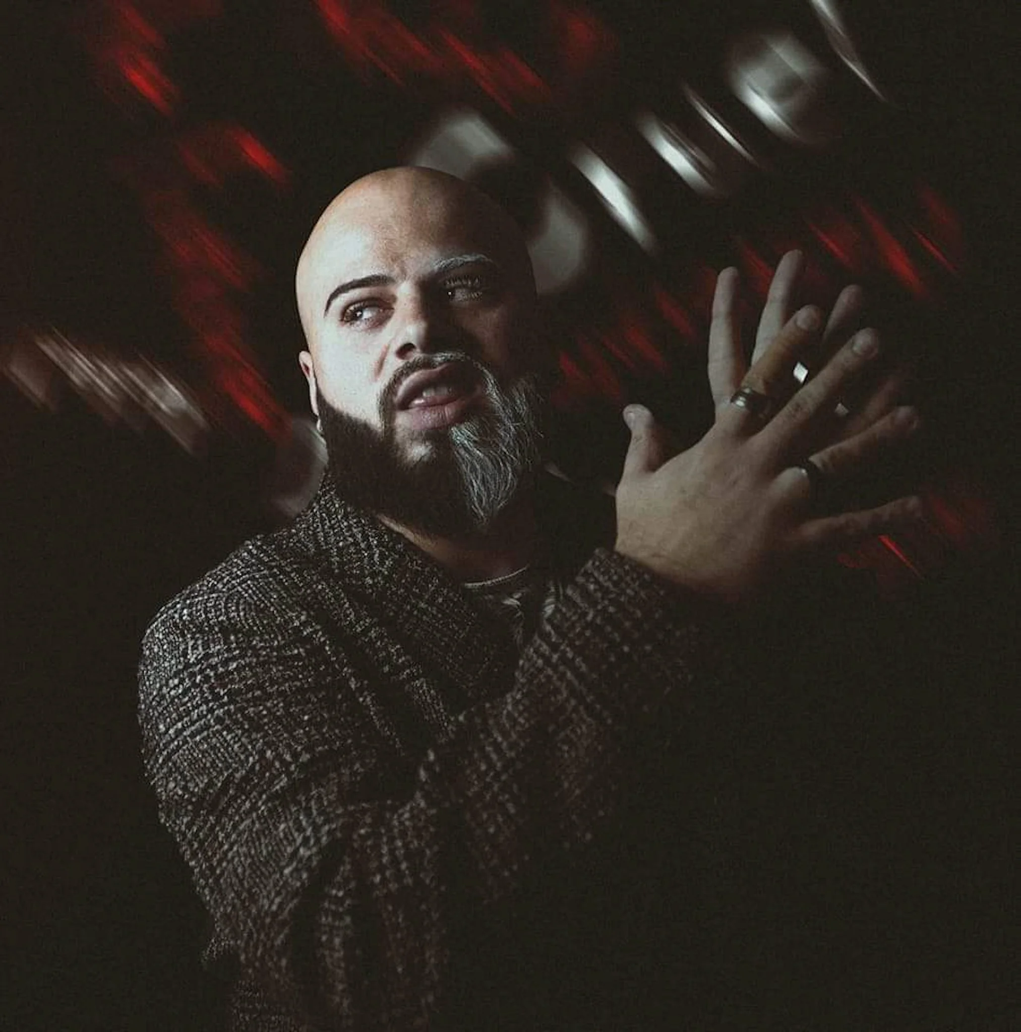 Close-up of a person with a bald head, light skin, and a beard, lying down in a dark environment with red and white blurry lights, and their hand raised near their face.