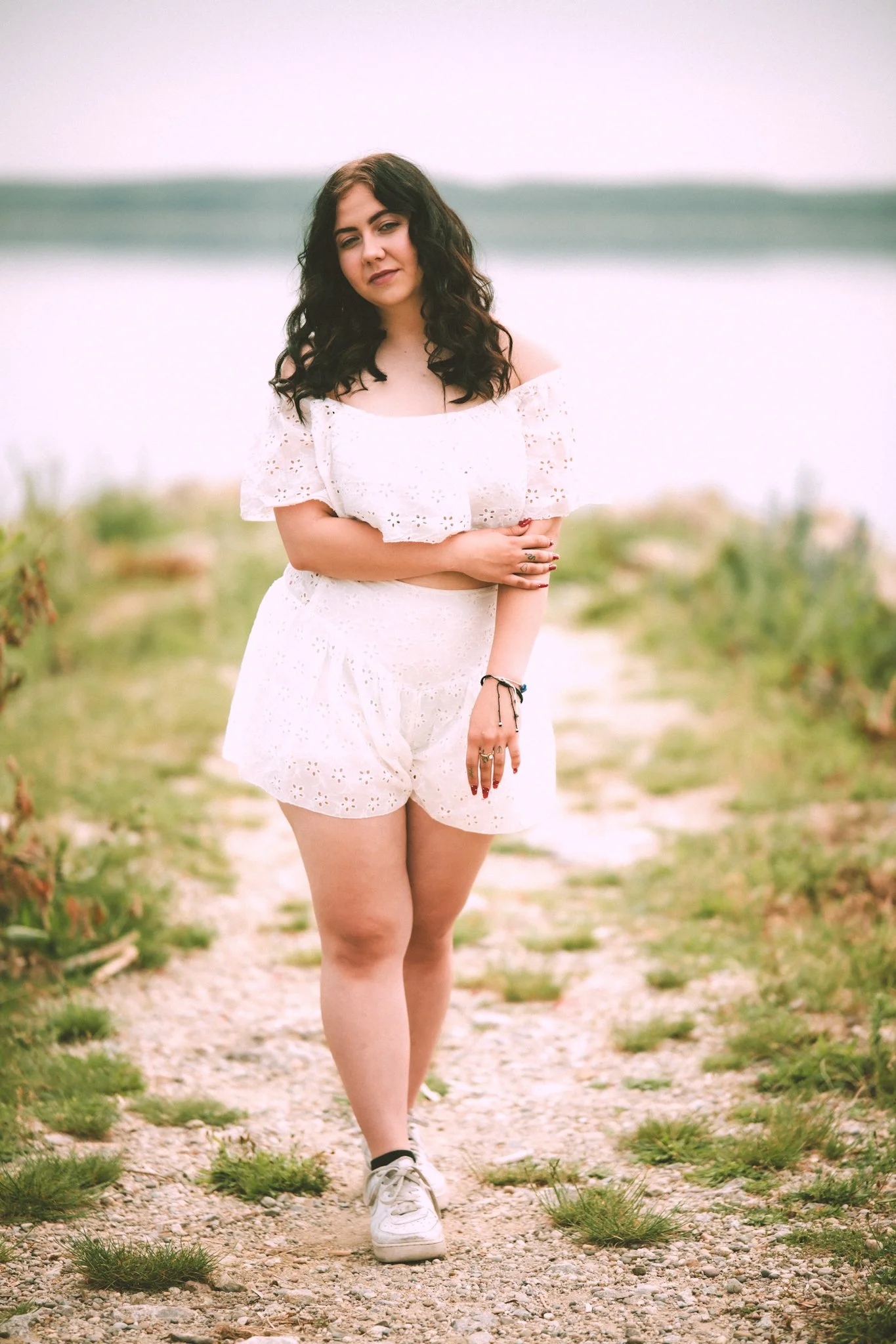 A young woman with dark curly hair, wearing a white eyelet off-shoulder dress and white sneakers, walking on a gravel path with small green plants, near a body of water under an overcast sky.