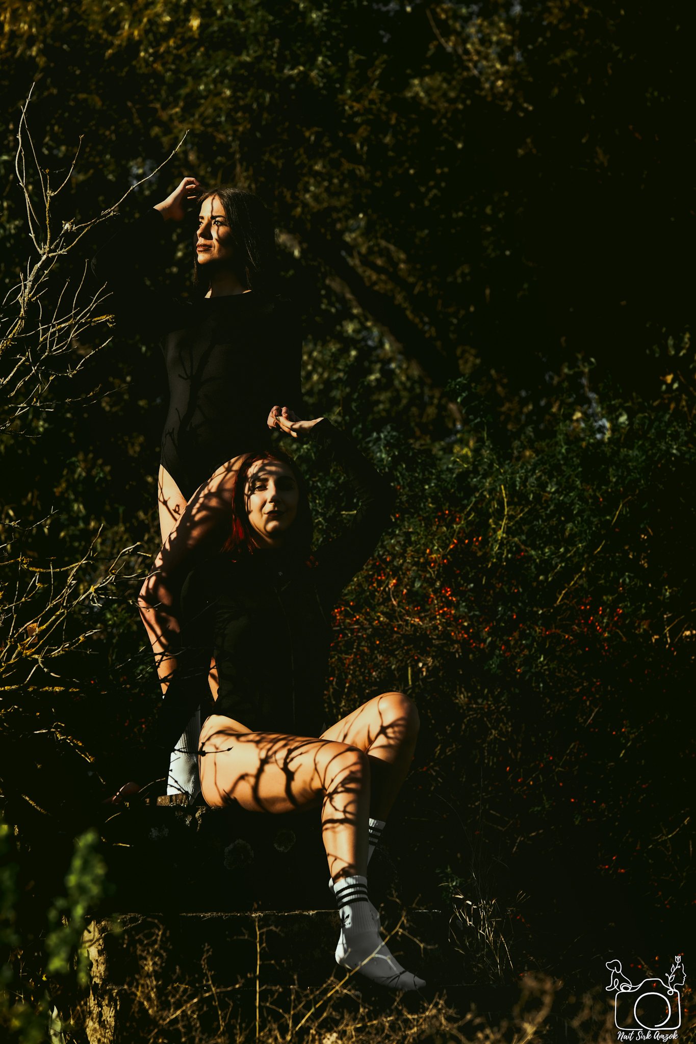 A woman standing on a tree branch in sunlight, casting shadows through the leaves, with arms raised, in an outdoor setting surrounded by trees.