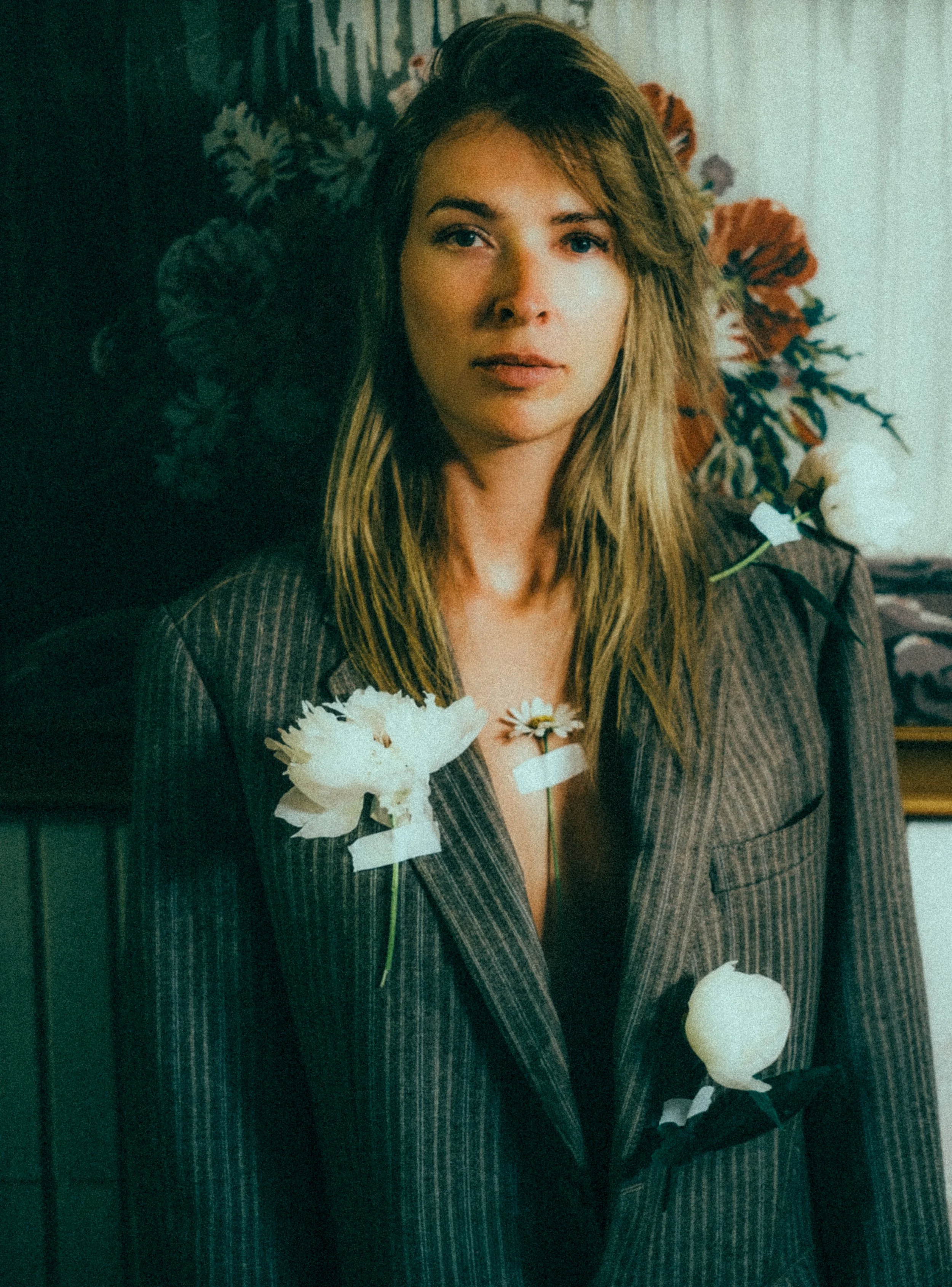 A woman with long, light brown hair and blue eyes, wearing a pinstripe suit, standing indoors near a dark wall with floral wallpaper. She has flowers taped to her chest and a small paper lantern hanging from her arm.