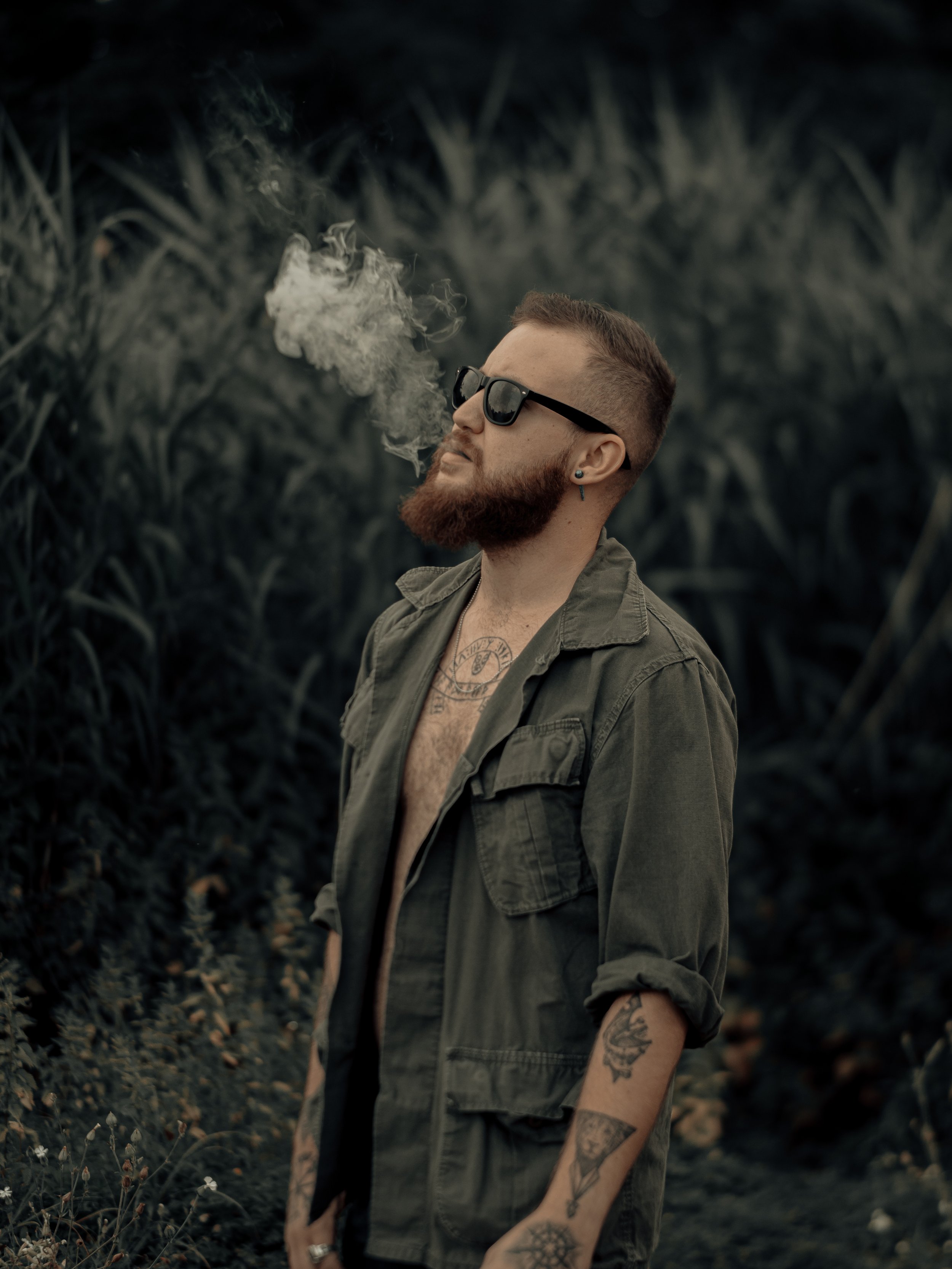 A man with a beard and tattoos, wearing sunglasses and an open green shirt, stands outdoors in a natural setting with dark foliage in the background, exhaling smoke.