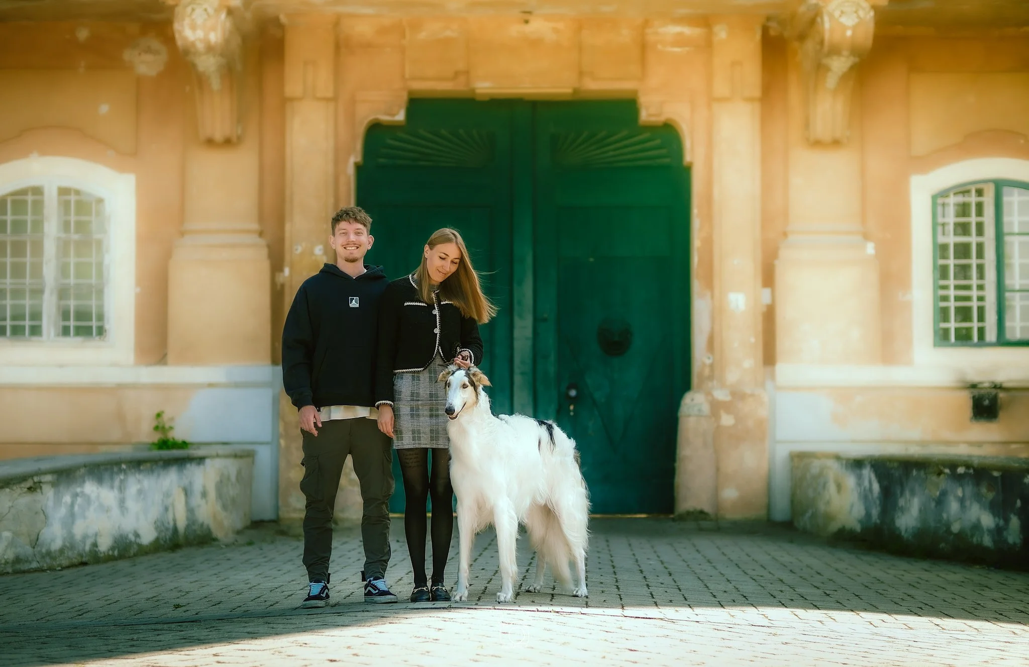 Two young adults, a man and a woman, standing outside a historic building with a large green door and ornate architecture, holding a white dog with black markings.