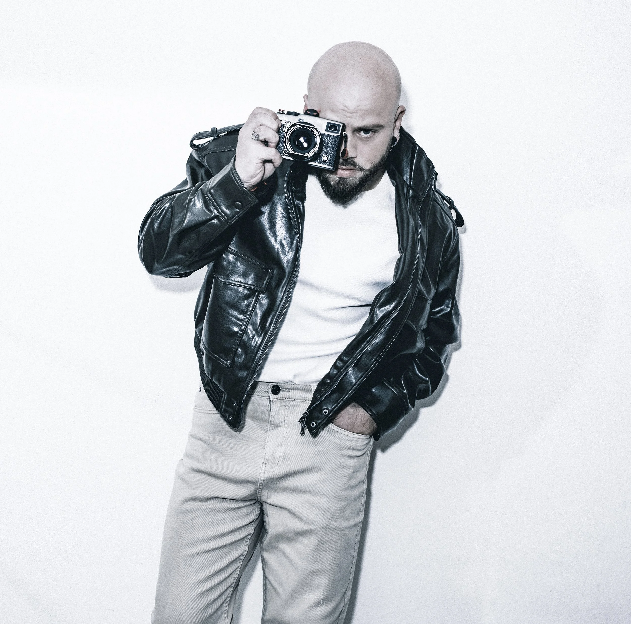 A man with a shaved head and beard is holding a vintage camera up to his eye, wearing a black leather jacket and light-colored pants against a plain white background.