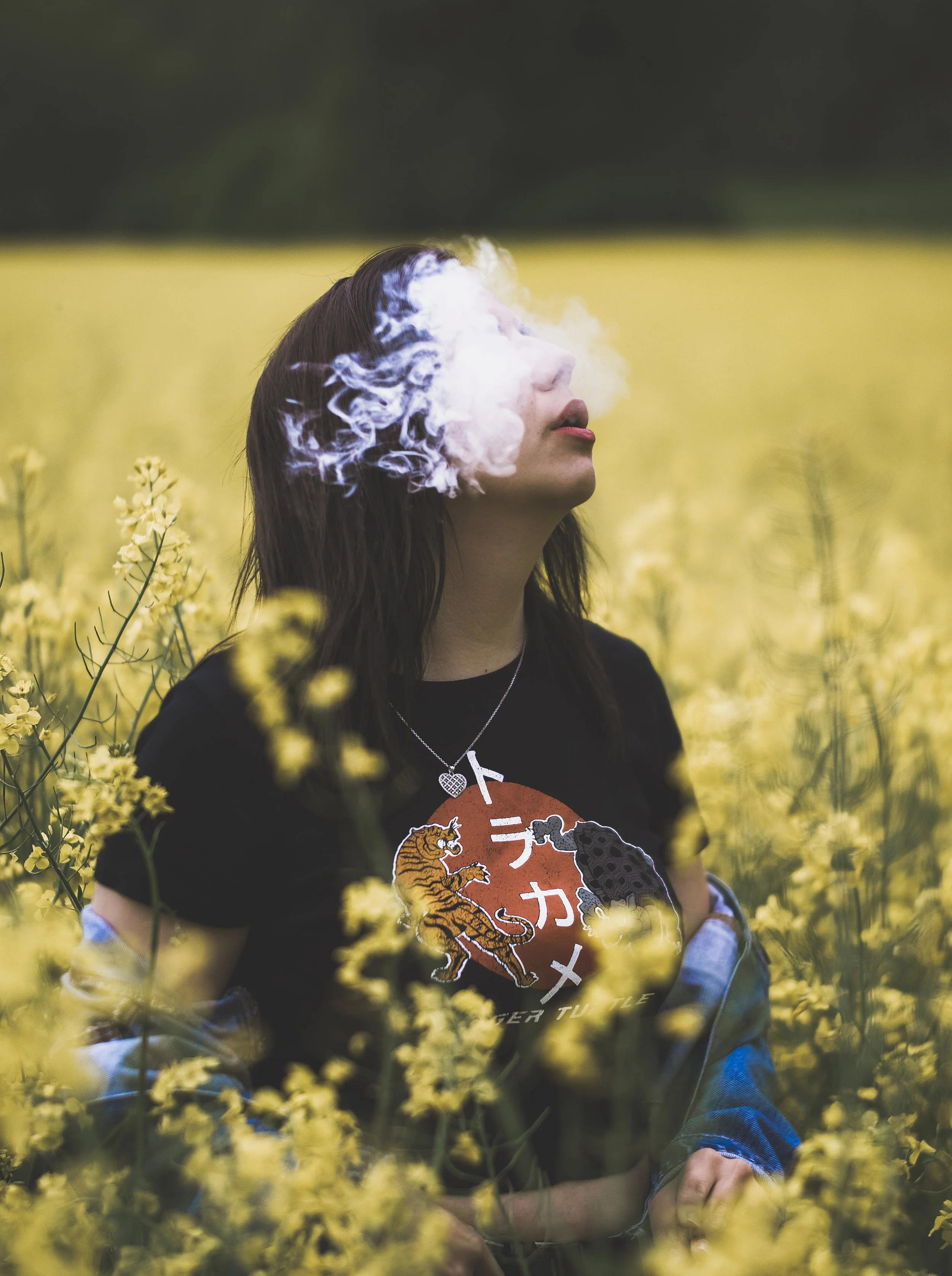 A woman with long dark hair and light skin is standing in a field of yellow flowers, with her head tilted upward and her face partly obscured by smoke or vapor near her face.