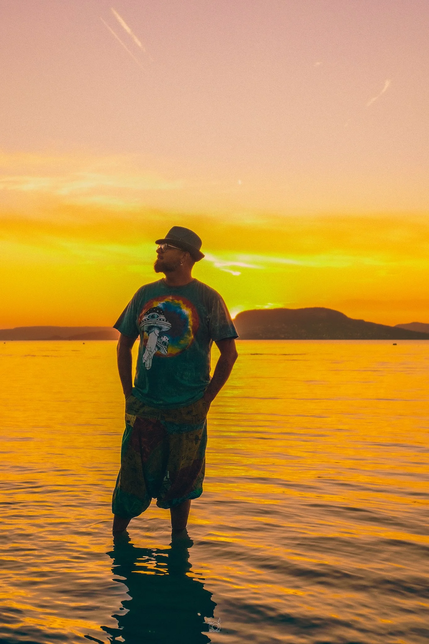 A man stands in shallow water during sunset, wearing a hat, glasses, a colorful T-shirt with a rainbow and a graphic design, and patterned shorts. The sky is orange and pink with some clouds and birds flying. The reflection of the sunset and the man 