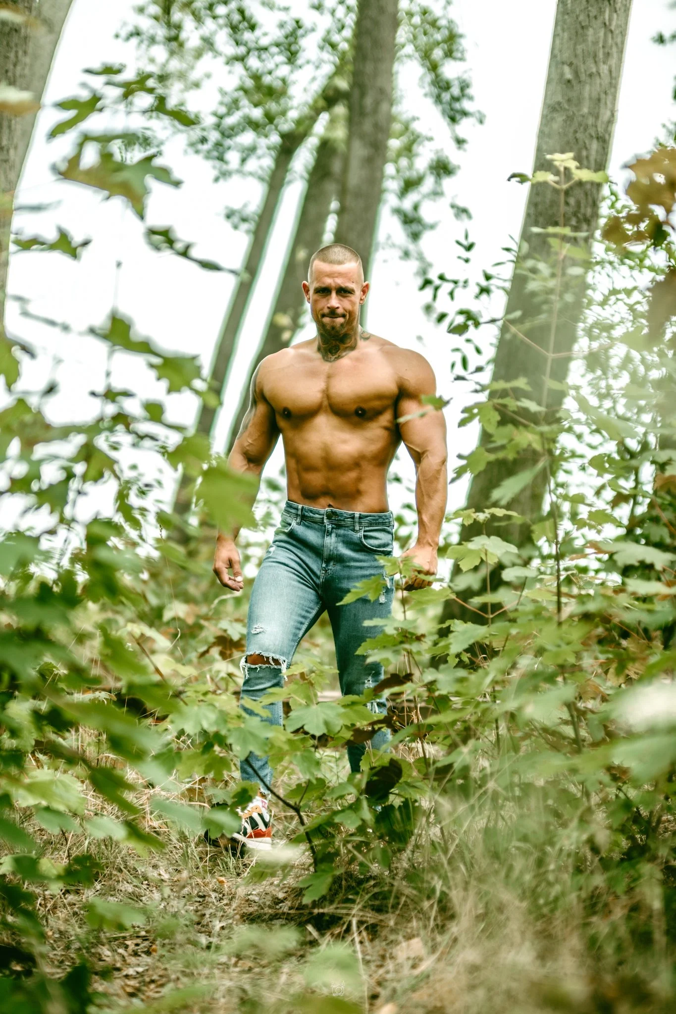 A shirtless man with tattoos and ripped jeans standing in a forest surrounded by green leaves and trees.