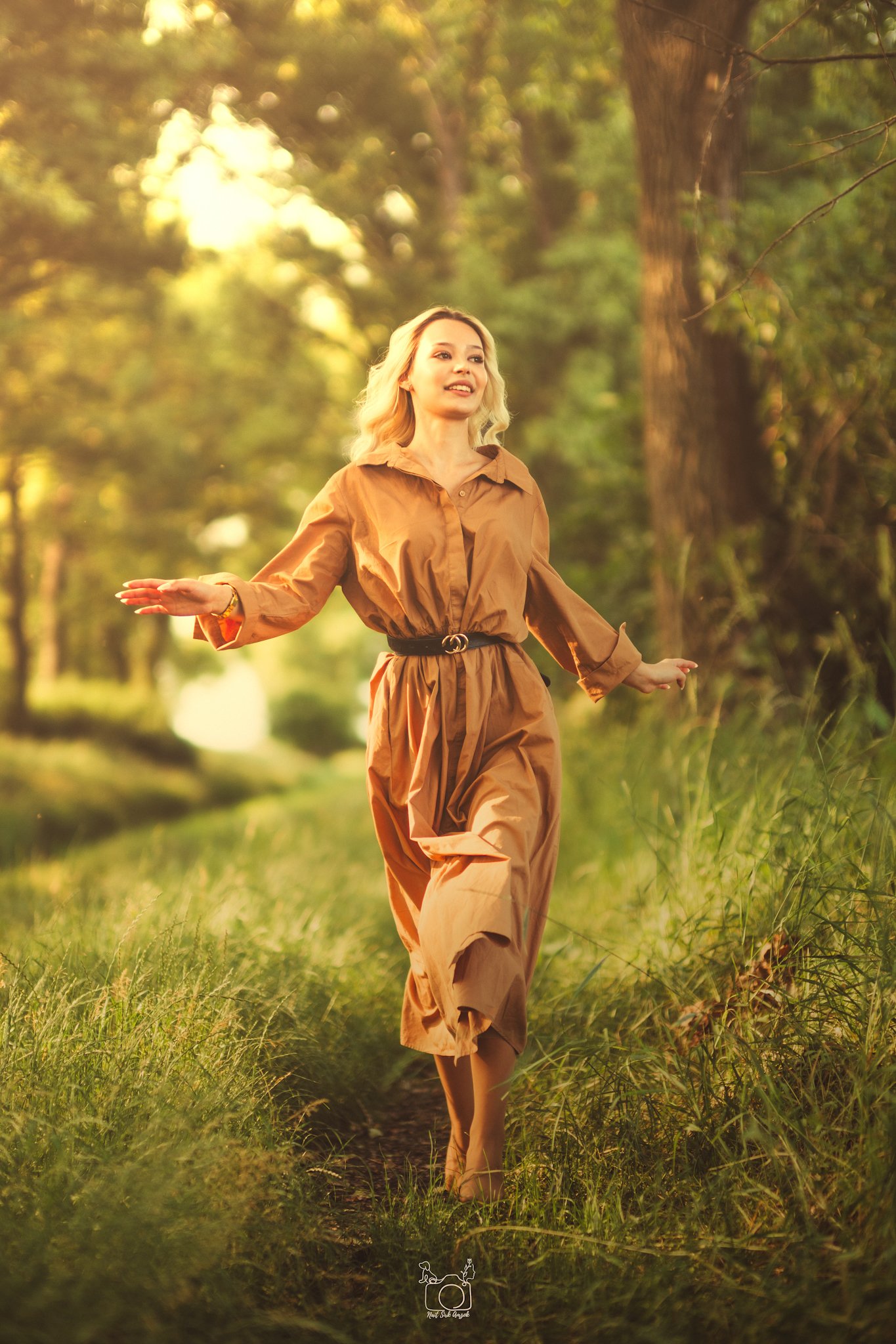 A woman with blonde curly hair smiling and walking in a sunlit forest trail during late afternoon, wearing a brown dress with a black belt.
