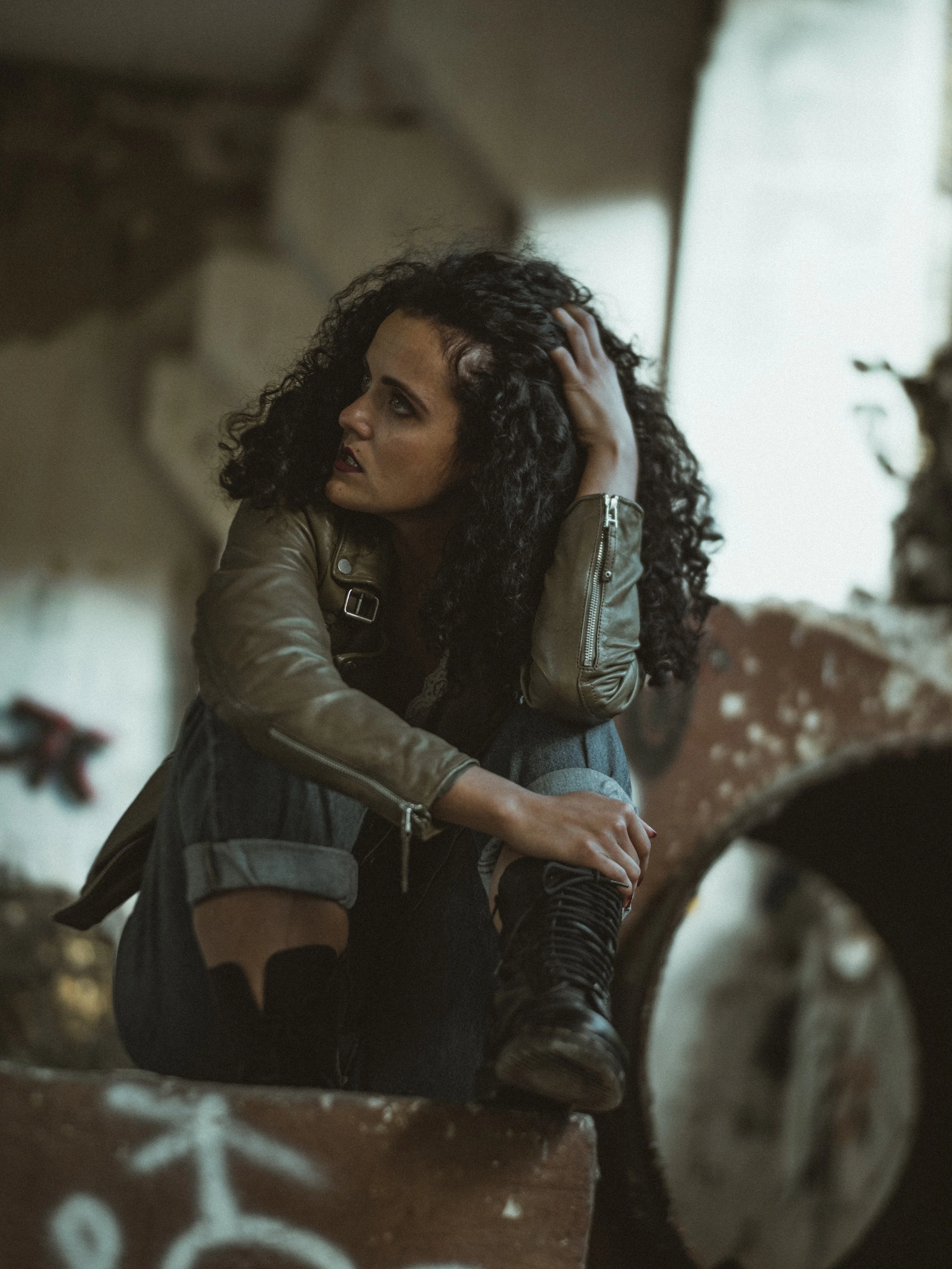 A woman with curly black hair and dark makeup sitting on a staircase, looking to her left, with her hand on her head. She is wearing a brown leather jacket, denim shorts, and lace-up boots.