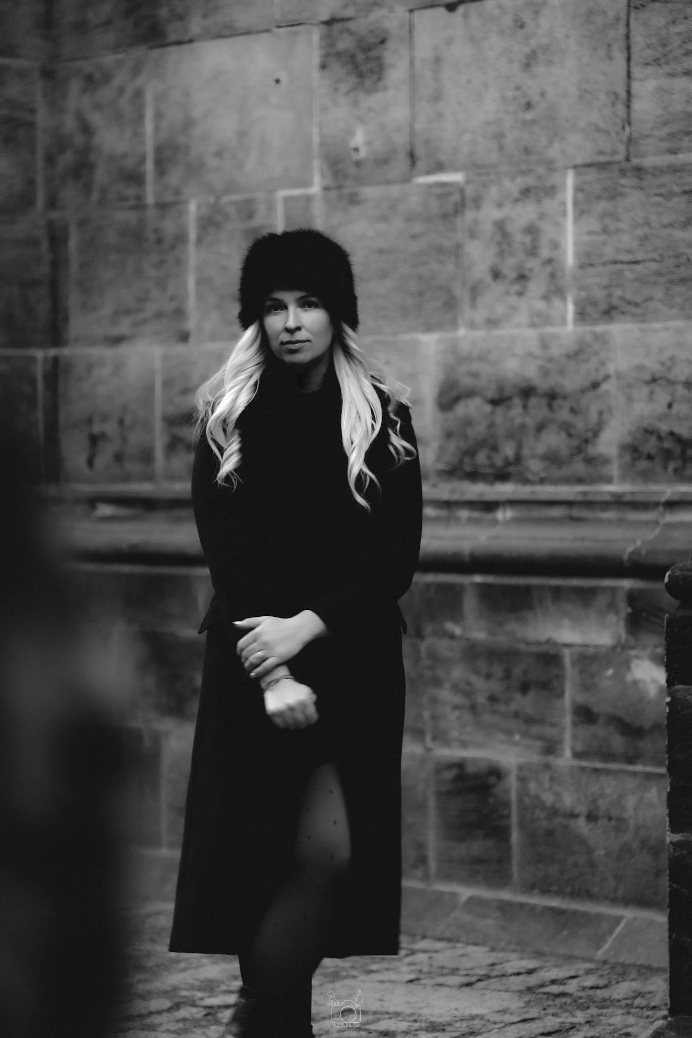 A woman with long, wavy blond hair wearing a black fur hat and black dress standing against a stone wall in a black and white photo.