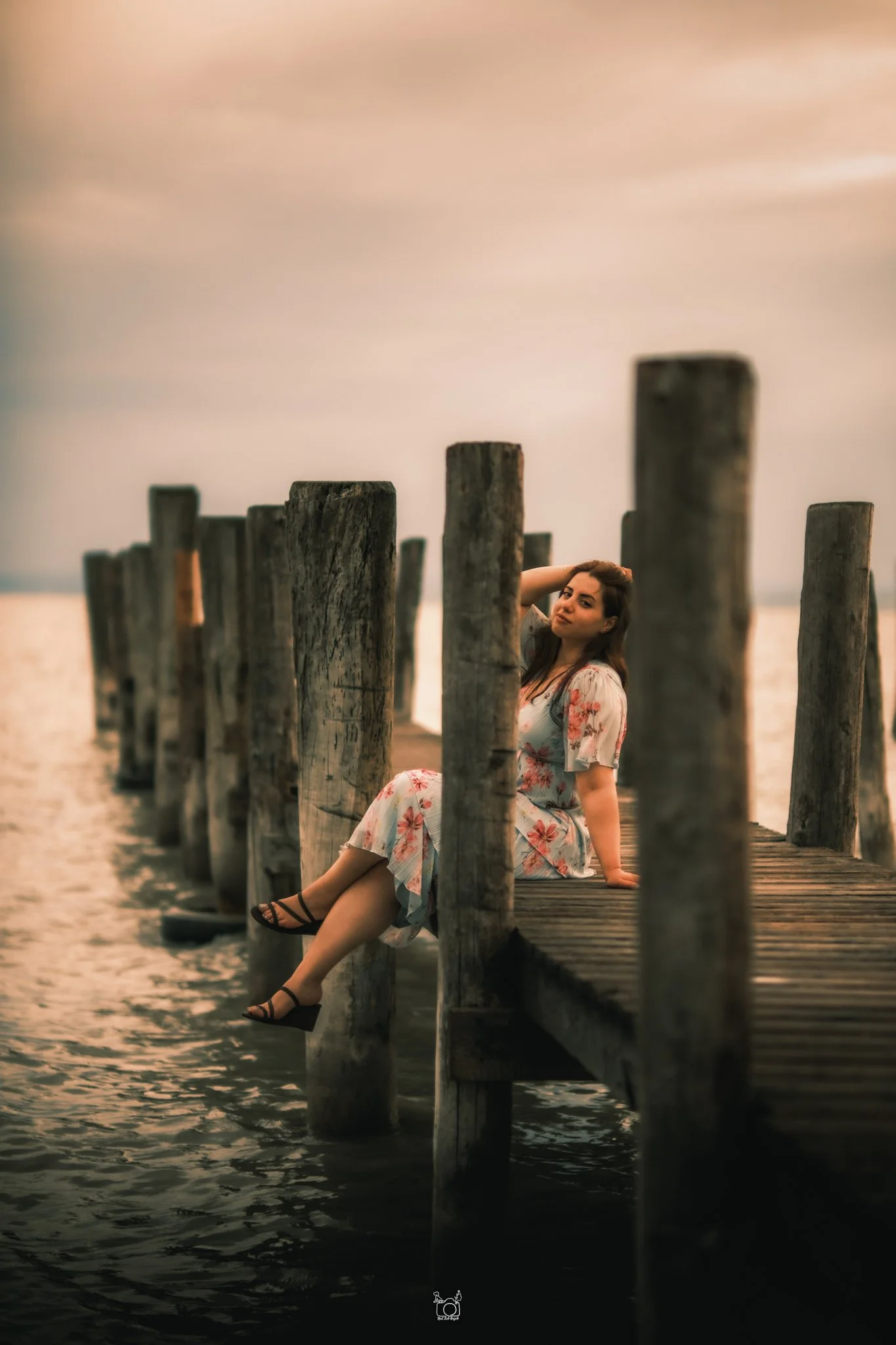 A woman sitting on a wooden dock by the water at sunset, wearing a floral dress and black sandals, with a row of weathered wooden posts behind her.