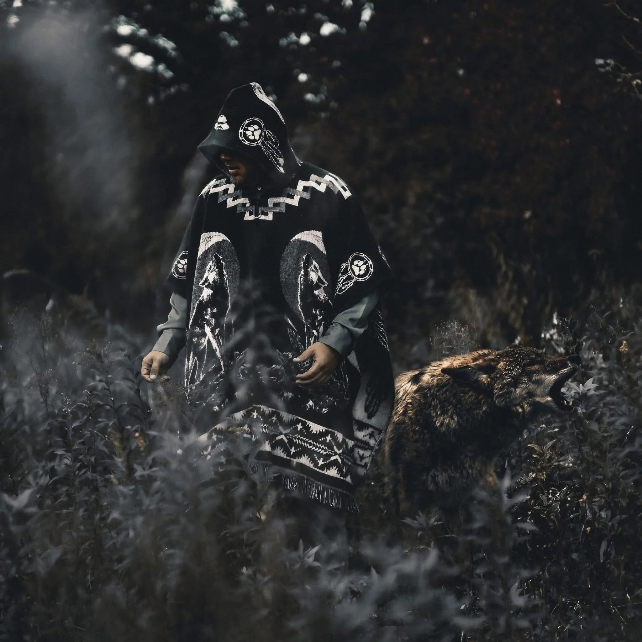 A young man with a beard wearing a black hoodie with white tribal and animal designs, sitting outdoors at night in a natural setting with trees and bushes.