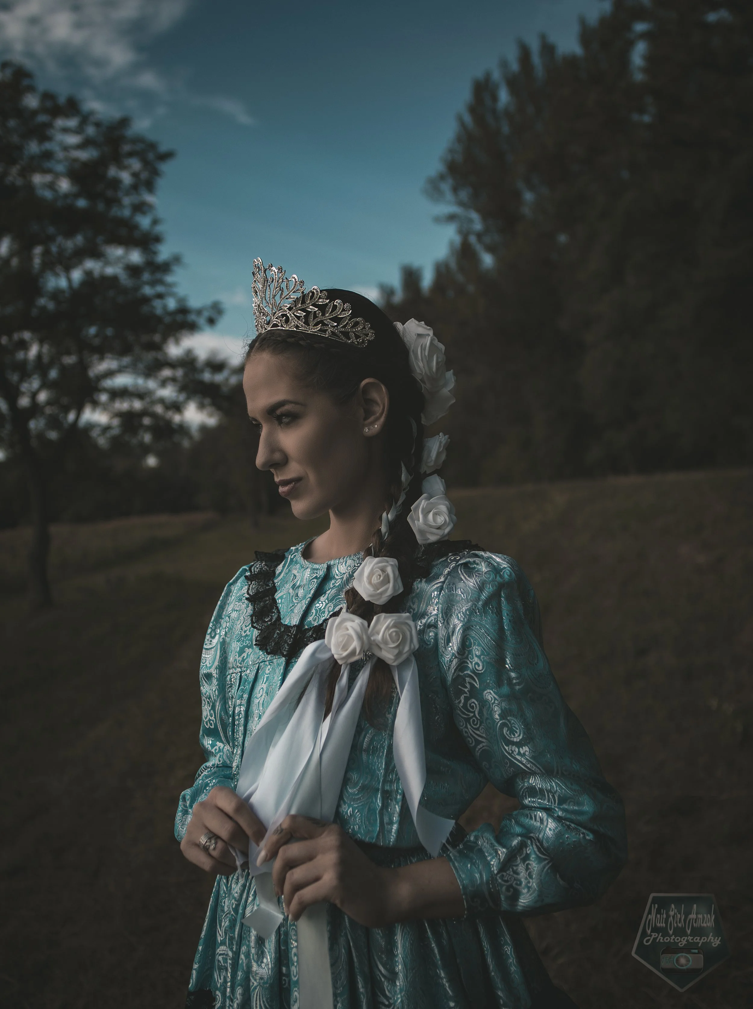 A woman in traditional Mexican dress with silver and white floral accessories, wearing a tiara, standing outdoors during dusk with trees and a cloudy sky in the background.