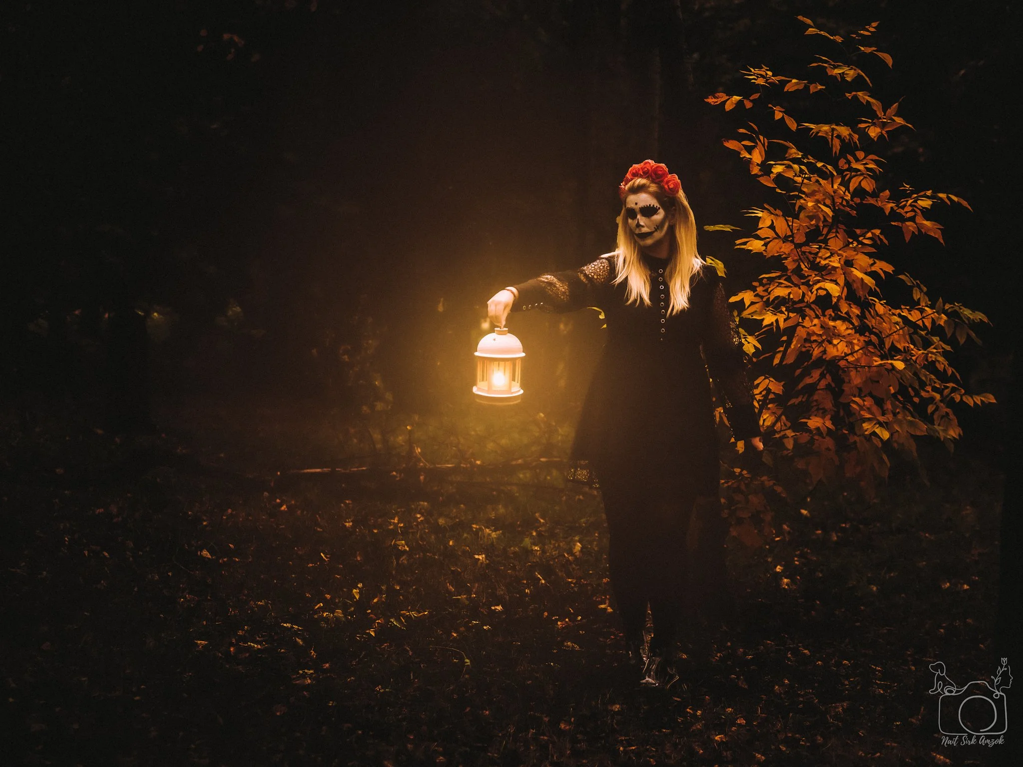 A person with face paint resembling a skeleton wearing a red flower crown, standing outdoors in a dark forest at night, holding a lantern emitting a warm glow, surrounded by autumn-colored leaves.