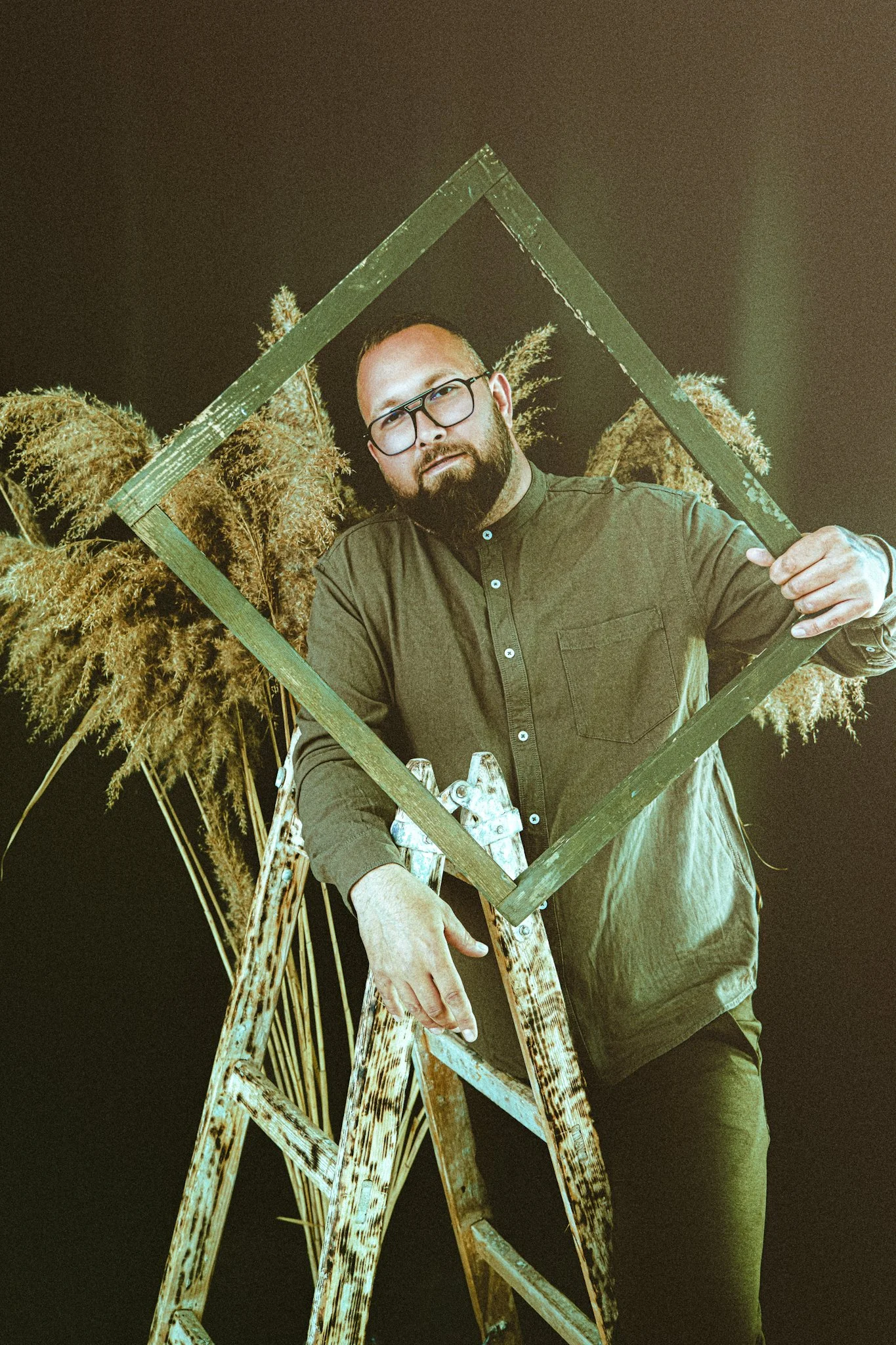 A man with glasses and a beard, wearing a green button-up shirt, holding a green wooden frame, standing in front of tall plants at night.