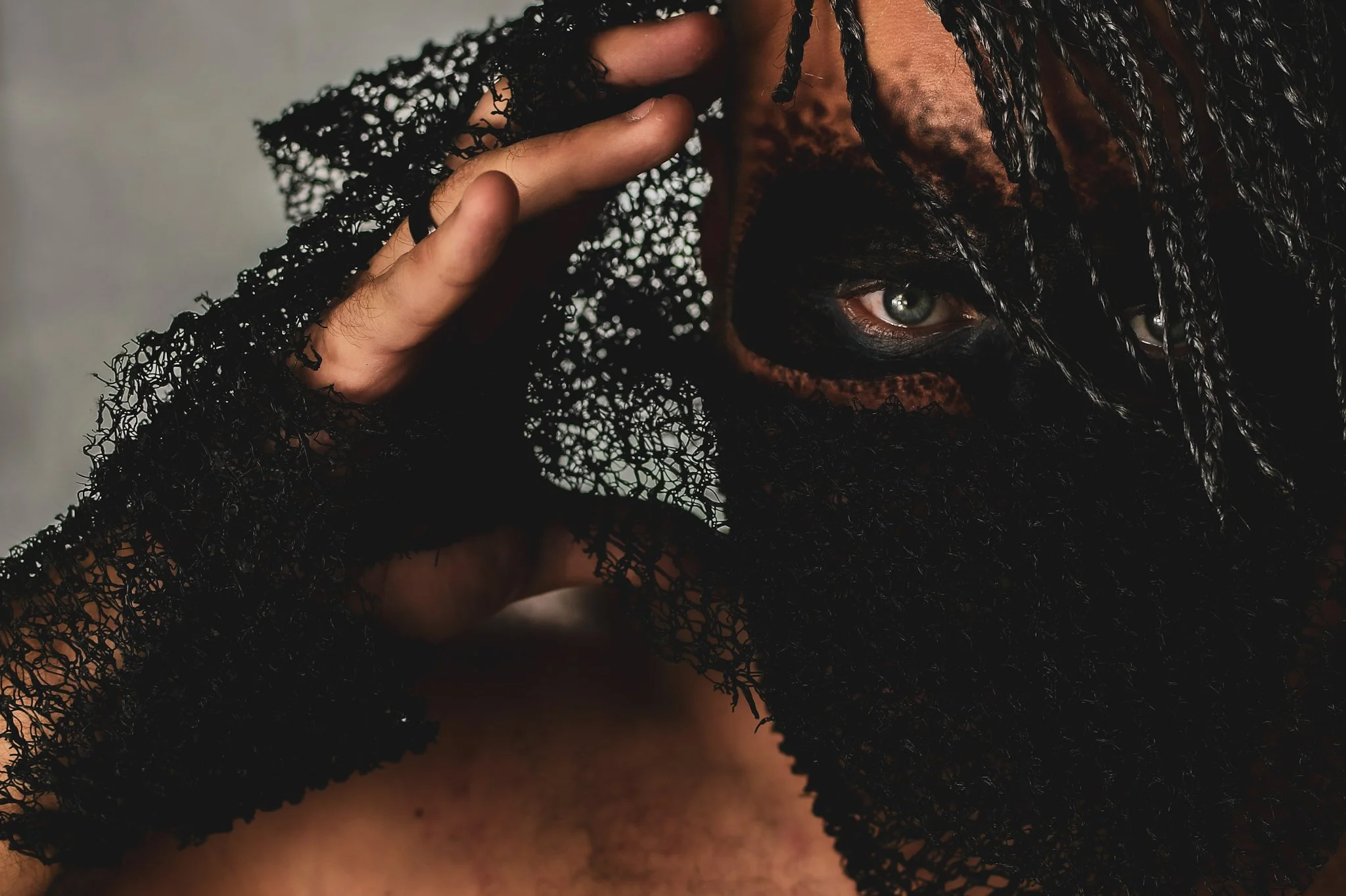 Close-up of a person's face partially covered by a black lace covering, revealing intense eyes and textured skin.