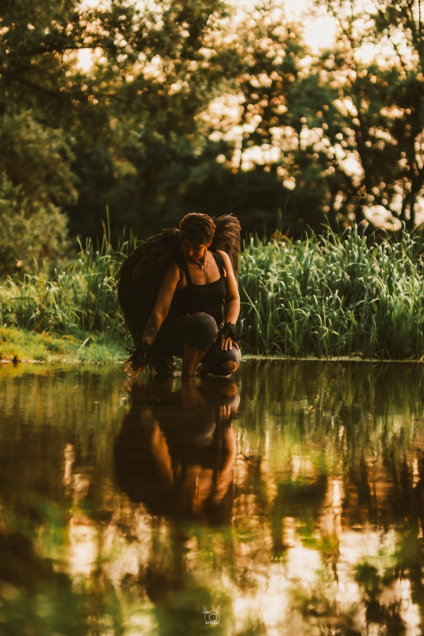 A person squatting in a water body during sunset, with tall grass and trees in the background.