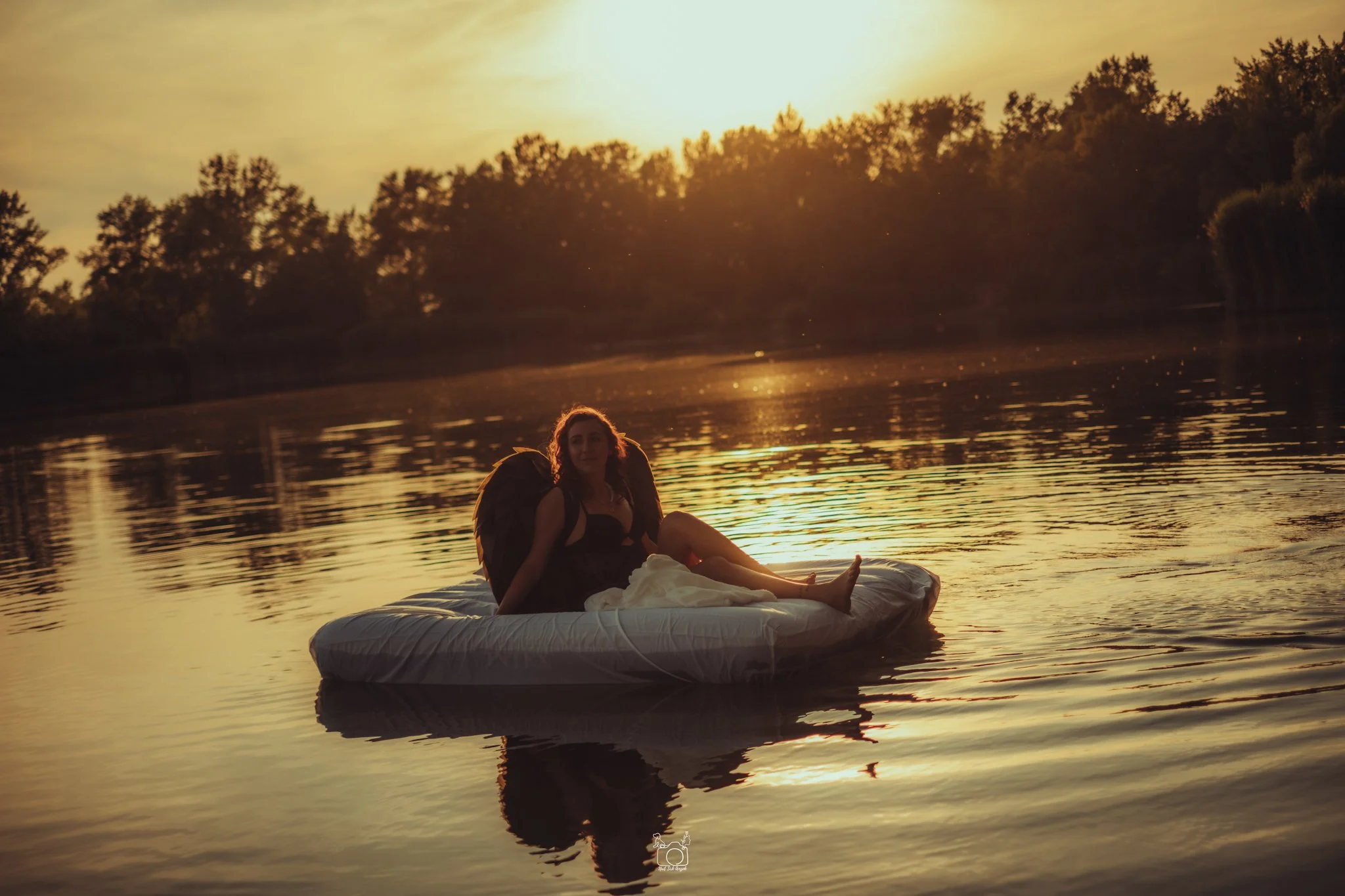 A woman with curly hair sitting on a raft on a calm river during sunset, wearing a black dress with brown feathered wings.