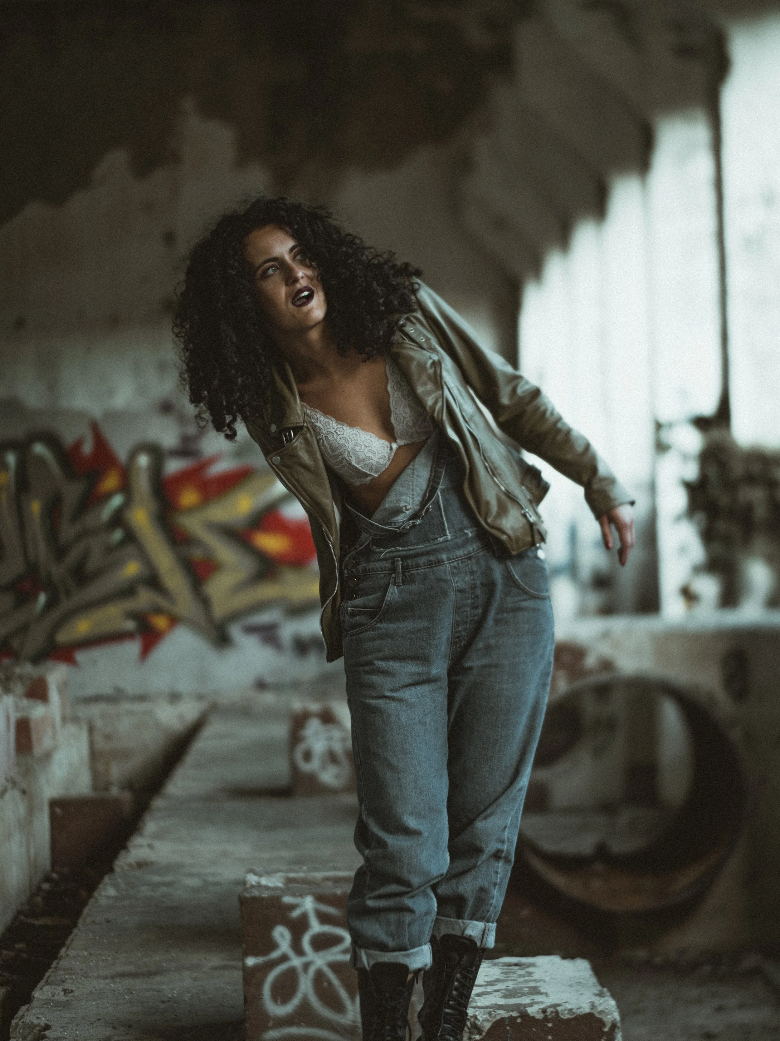 Woman with curly hair wearing a leather jacket, lace top, and jeans standing indoors near graffiti-covered wall.