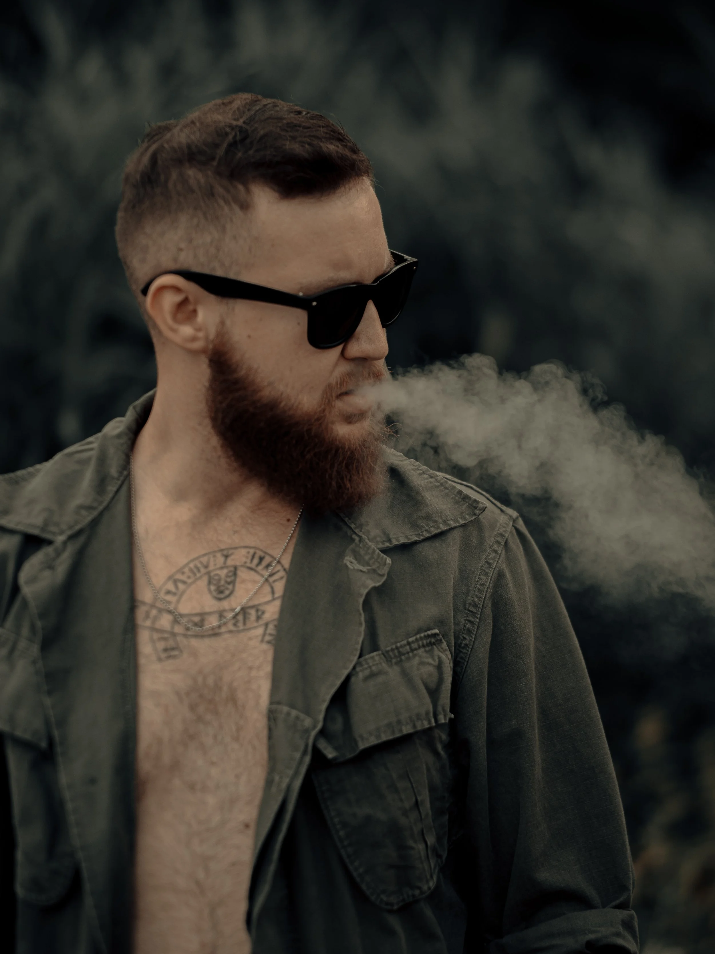 A man with a beard and short hair wearing sunglasses, a tattoo on his chest, and an unbuttoned green jacket, exhaling smoke outdoors.