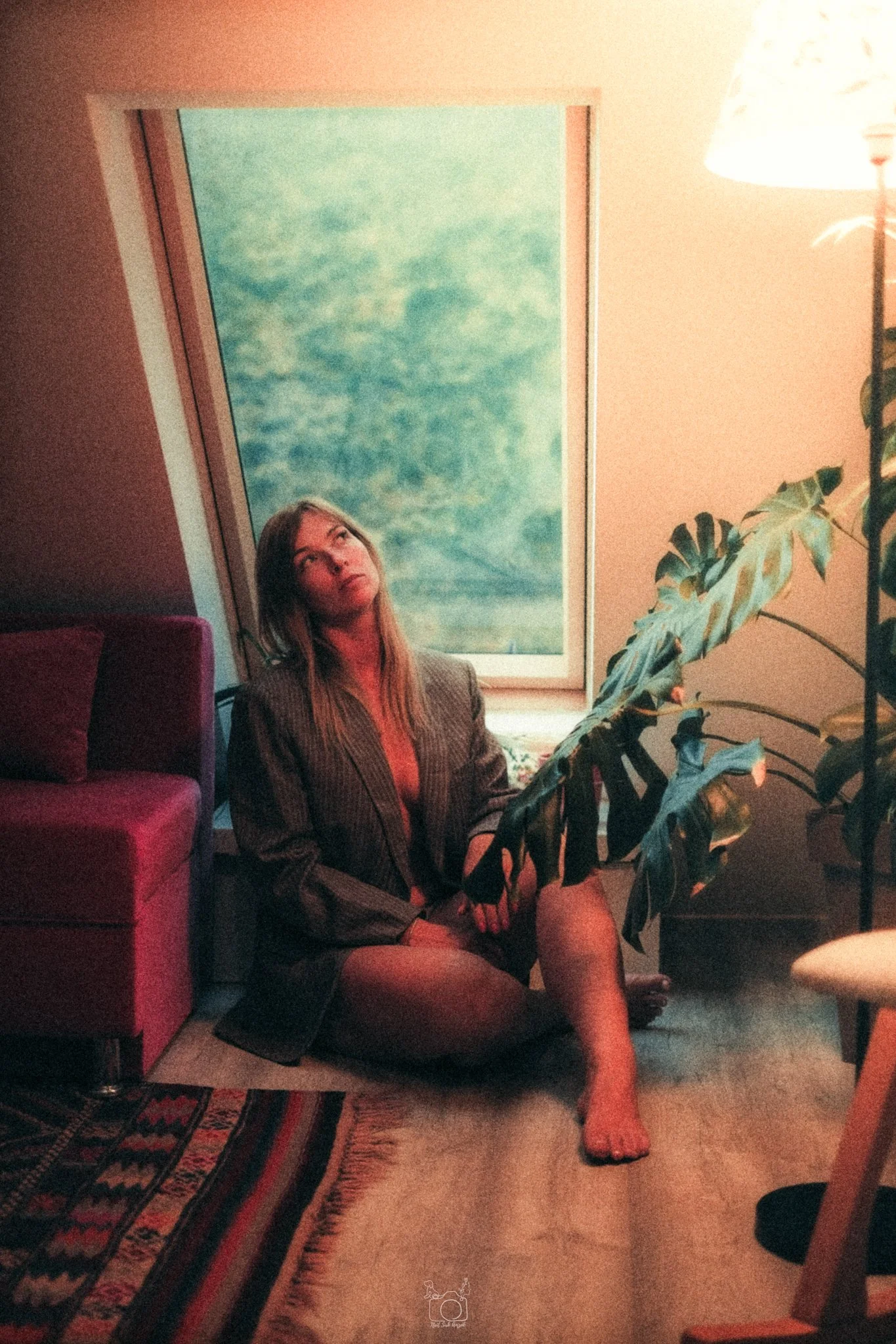 A woman with long hair, dressed in a blazer and shorts, sitting on the floor next to a large green houseplant and a window with a dreamy sky scene, with a lamp and furniture around