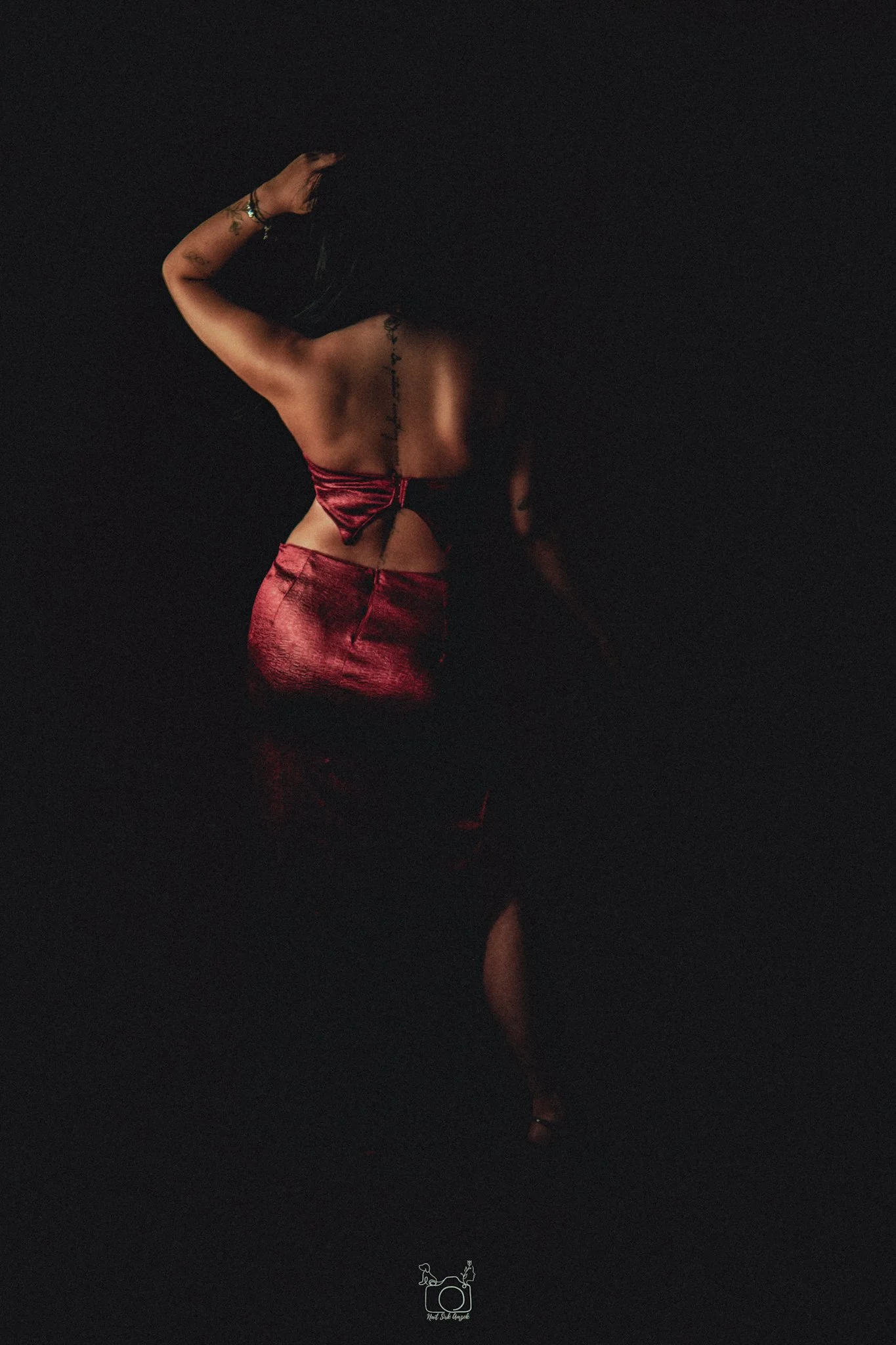 A woman standing in a dark environment illuminated by a small light, wearing a red metallic dress, with tattoos on her arm and back, and holding a black hat or similar object above her head.
