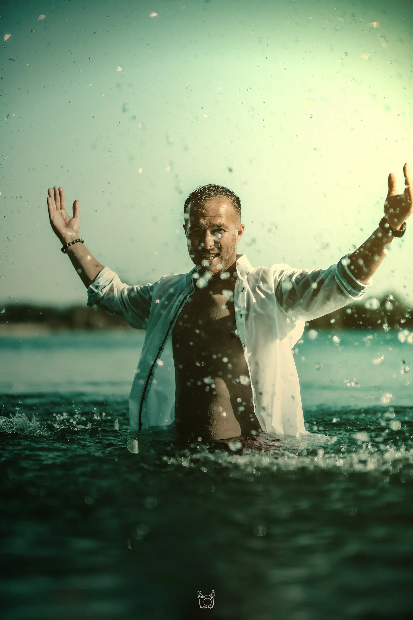 A man with water on his face and wet hair standing in the water at the beach with arms outstretched, wearing an open white shirt and smiling.