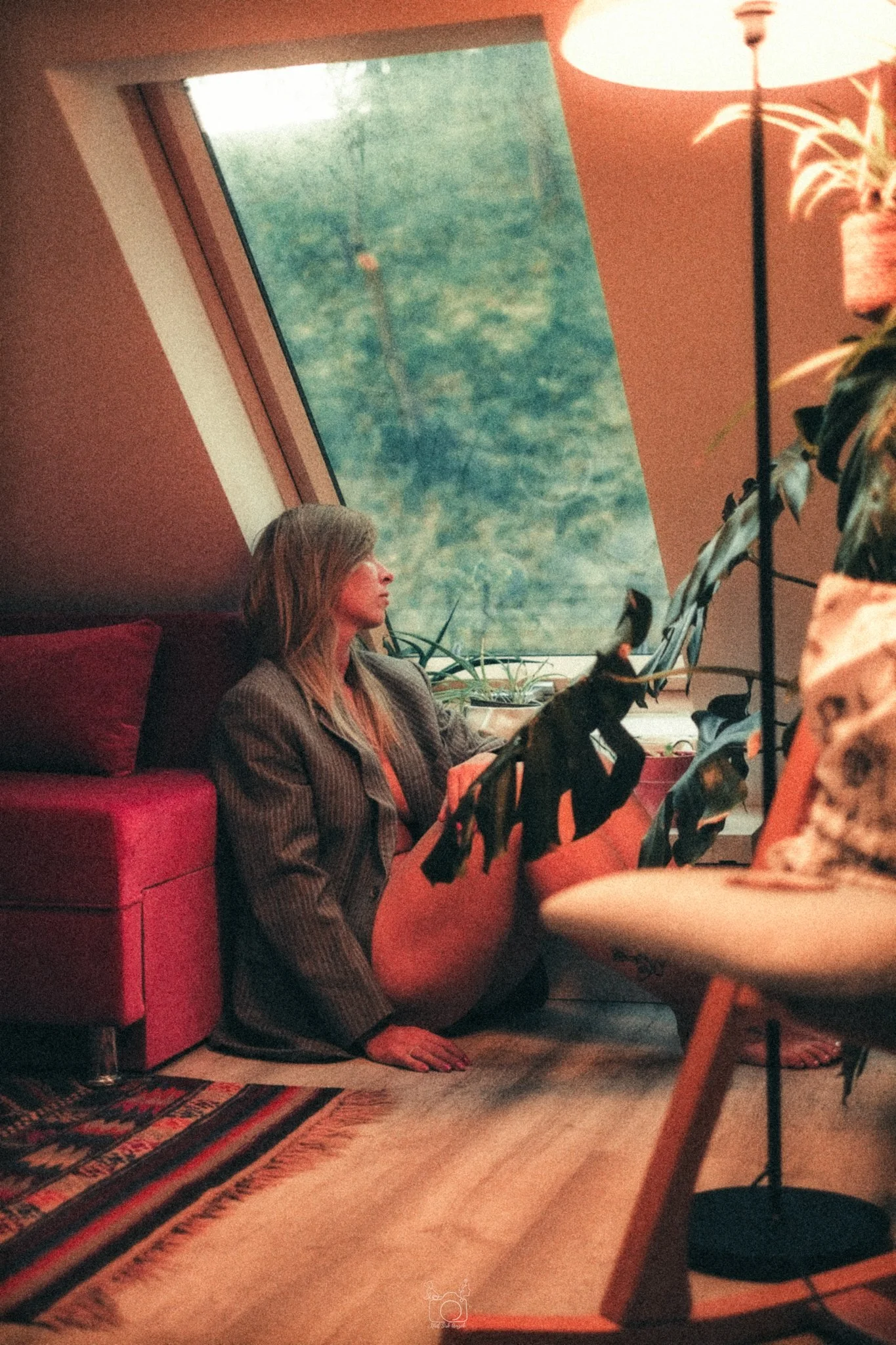 A woman with long, gray hair sitting on the floor next to a red sofa in a cozy room, with a large window showing green trees outside. She is wearing a striped jacket and appears to be relaxing or deep in thought. The room has warm lighting, potted pl