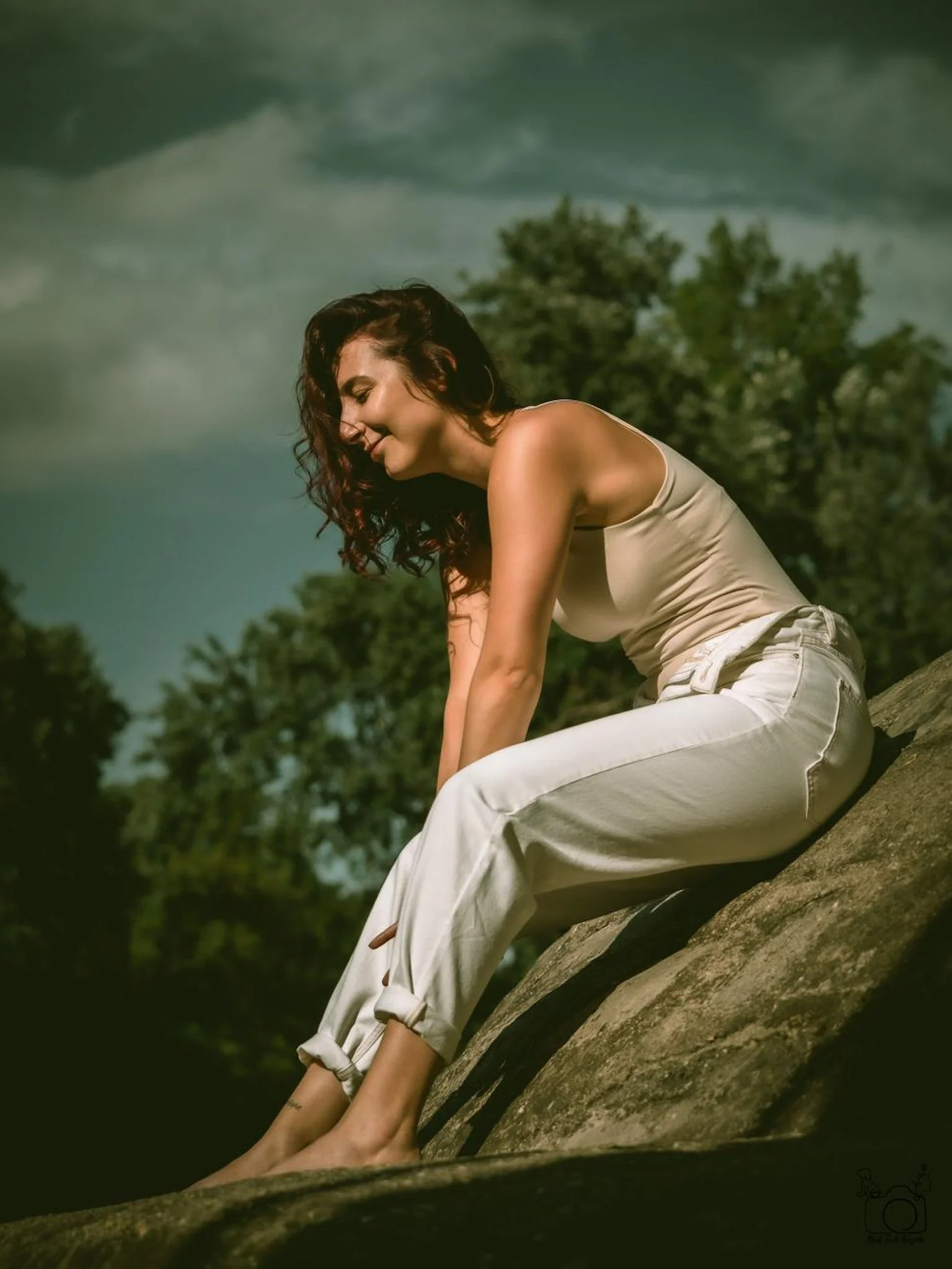 A woman with dark, curly hair in a sleeveless beige top and white pants, sitting on a large tree log outdoors, with a background of green trees and a cloudy sky.