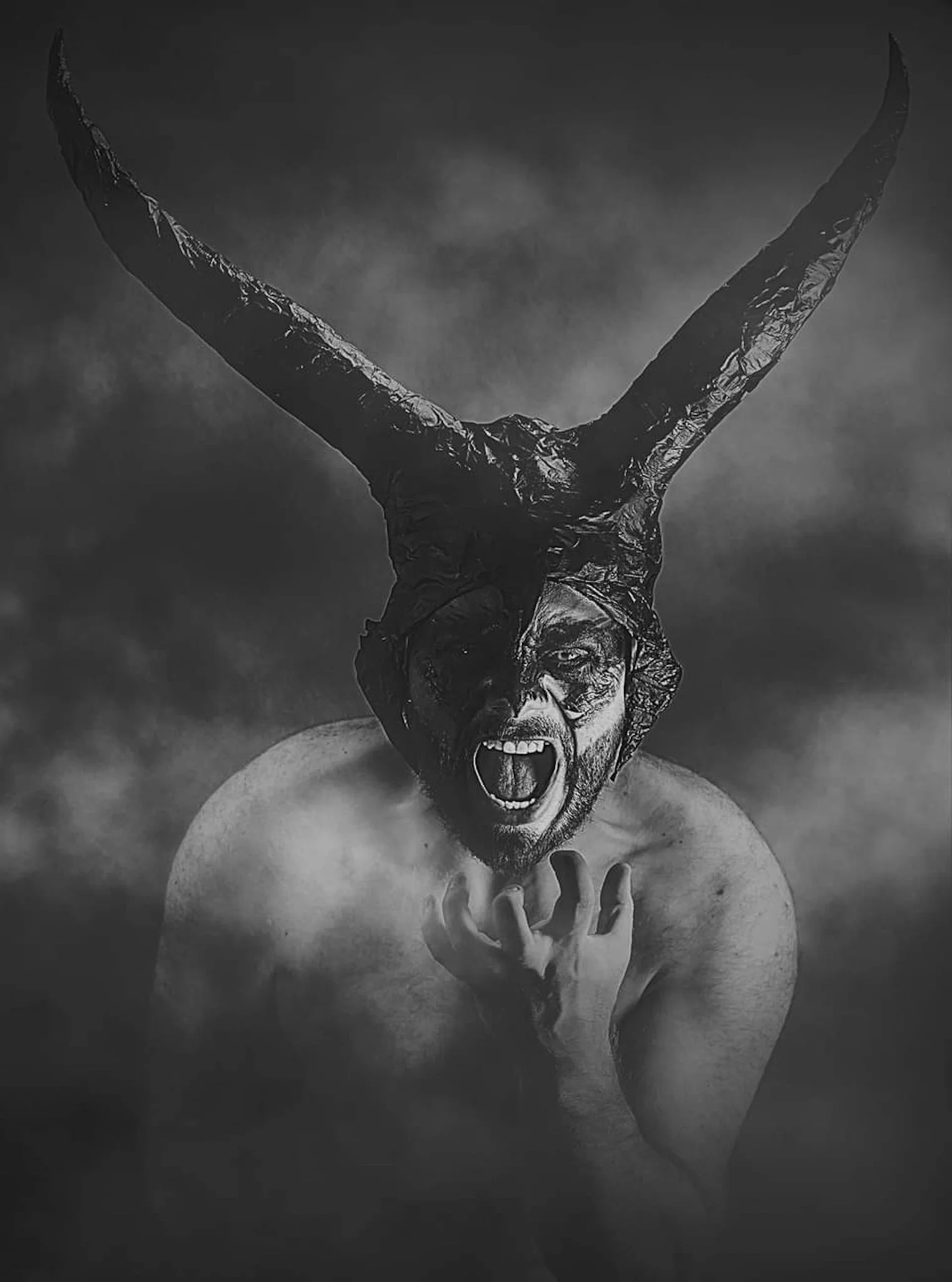 Black and white photo of a man with a beard, glasses, and curly hair, shouting with his mouth open and one hand on his chest, with a sculpture of a horned animal's head on his head.