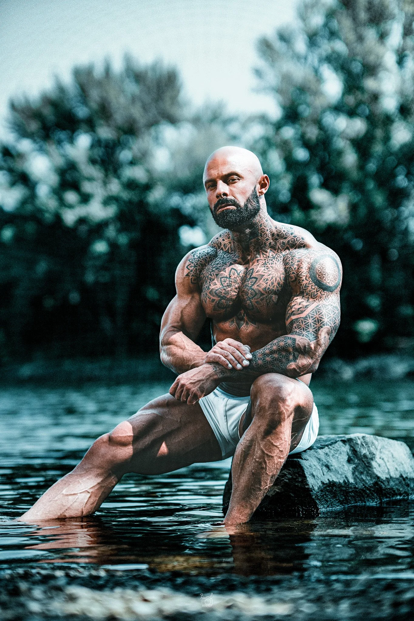 A bald, tattooed man with a beard kneeling on a rock in a body of water, with his arms crossed over his knee and looking at the camera. The background features blurred trees and a cloudy sky.