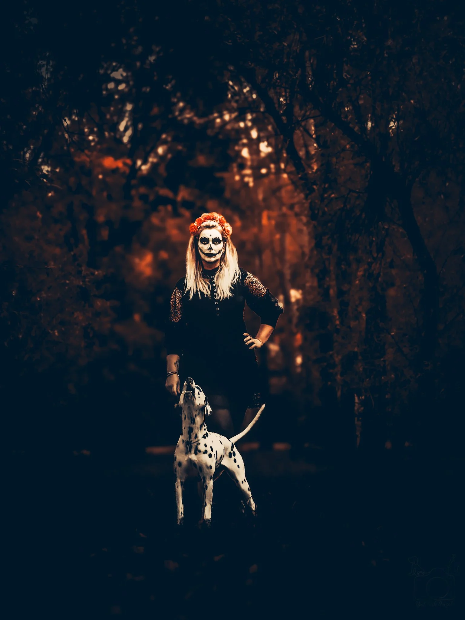 A person with face painted in a Day of the Dead style, wearing a flower crown, standing outdoors in a wooded area at dusk with warm lighting. They are holding a Dalmatian dog on a leash.