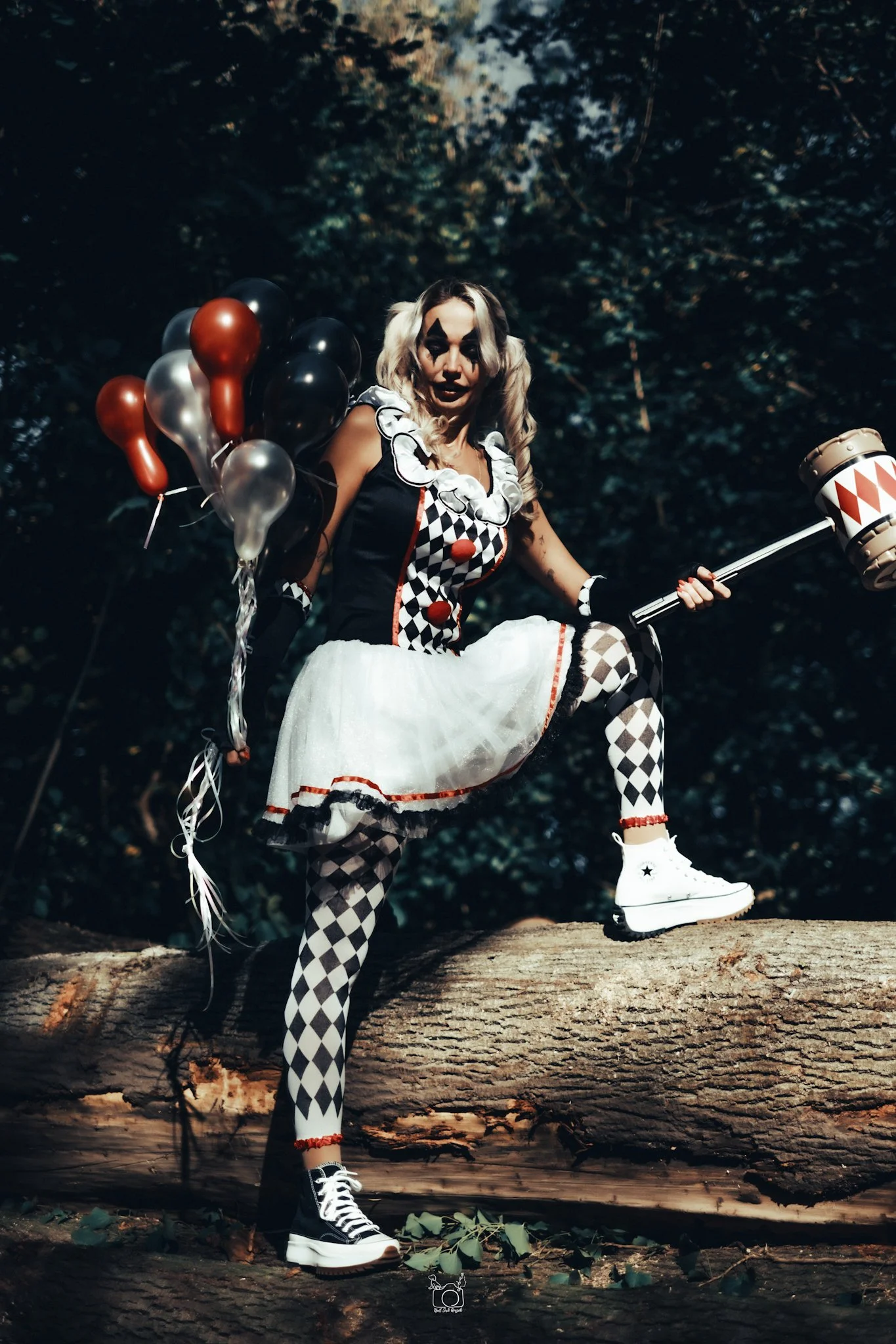 A woman dressed as a clown with black and white checkered tights, a black and white clown costume, and a white tutu, standing on a log in a dark forest. She holds a balloon bouquet and a mallet, with her face painted like a clown, and has blonde hair